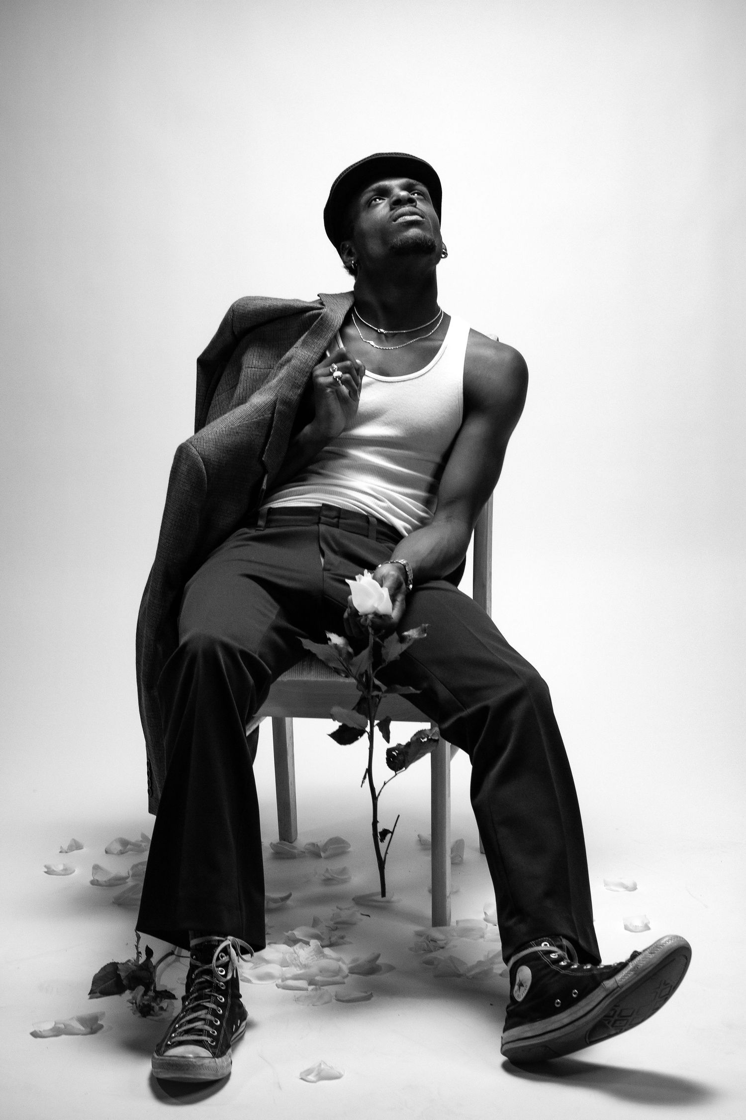Black and white photo of a young man sitting on a chair holding a rose, wearing a tank top and Converse sneakers.