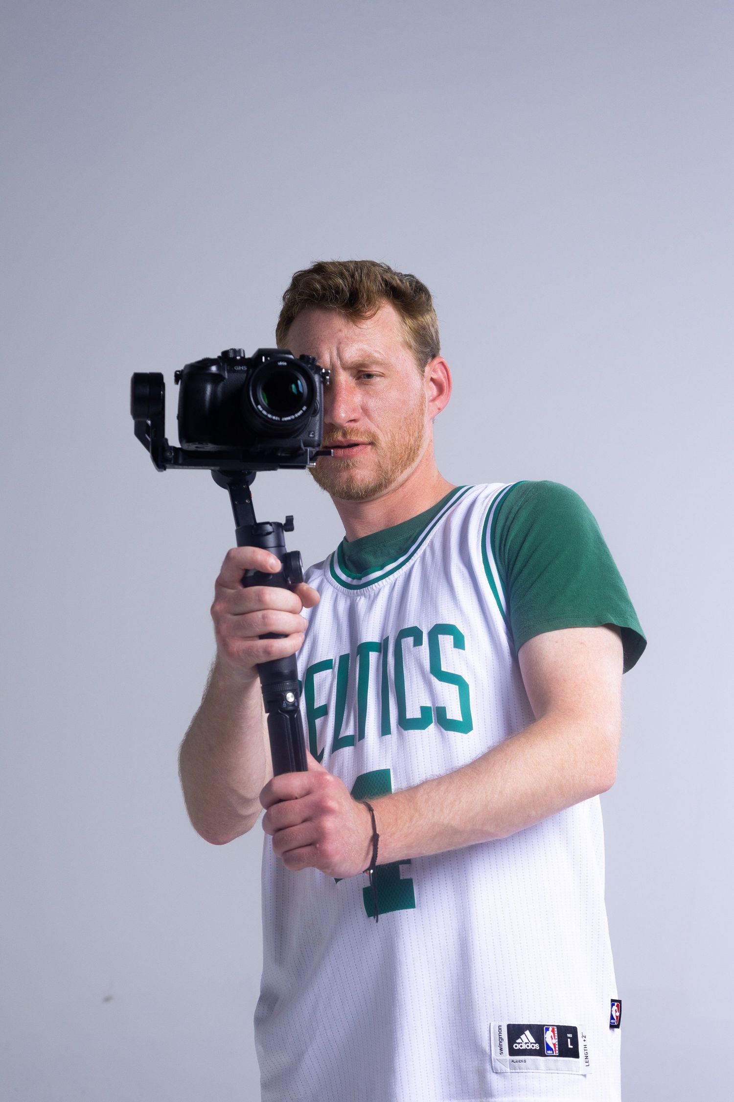 Man in Celtics jersey holds camera on gimbal stabilizer against gray background.