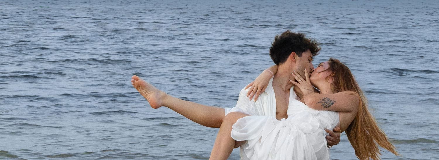 A couple in white outfits embrace and kiss while standing in front of a serene ocean backdrop.