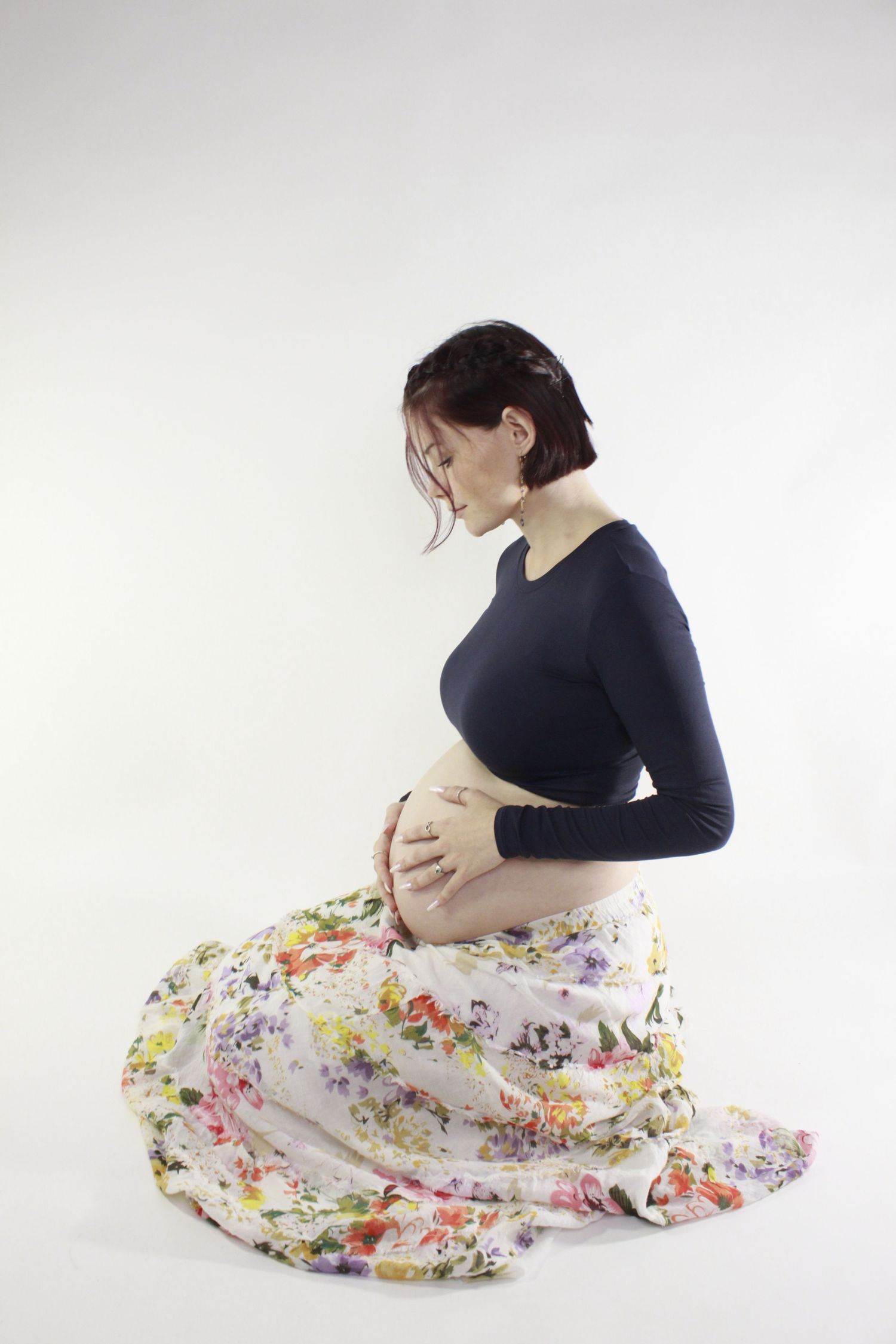 A pregnant woman in a navy top and floral skirt kneeling while cradling her baby bump on a white background.