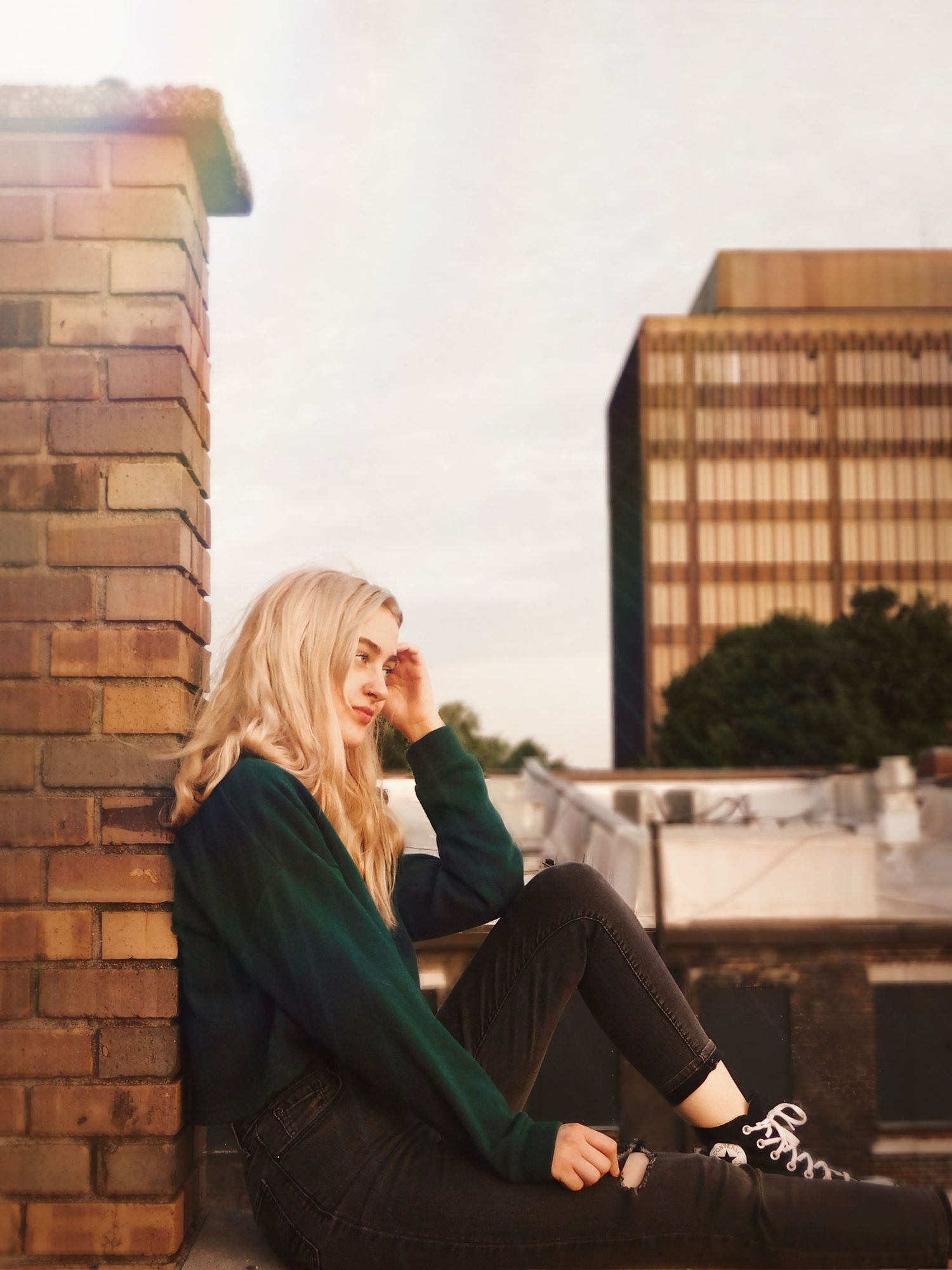A blonde woman sits against a brick wall on a rooftop, wearing a green sweater and black jeans, in urban setting.