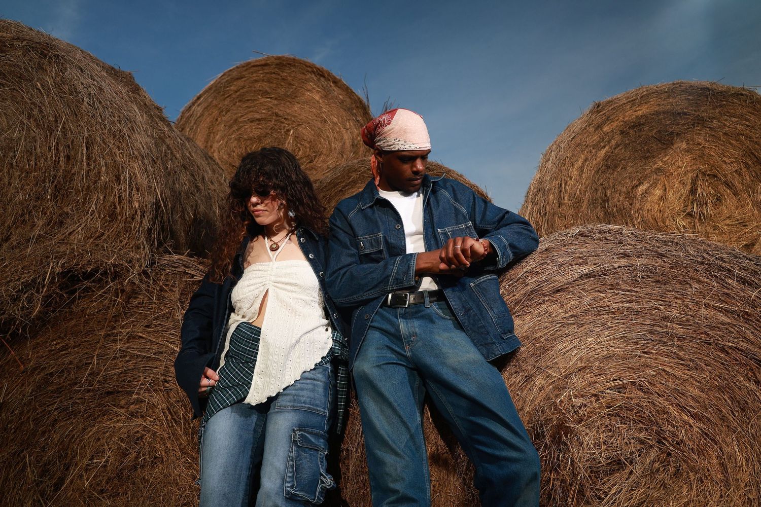 Two people in denim outfits pose against large hay bales under a blue sky in a stylish rural fashion shoot.