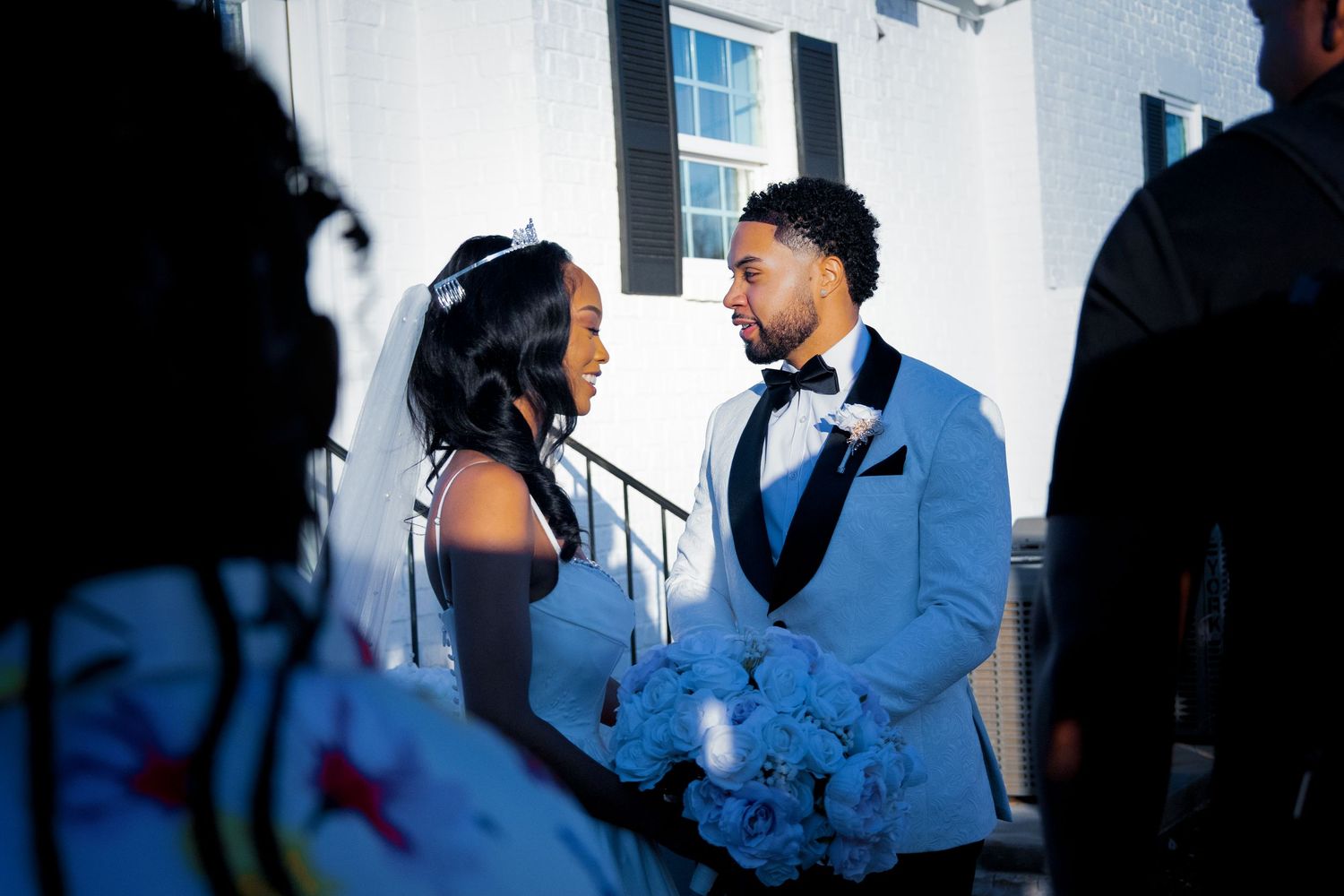 A bride and groom share a romantic moment outdoors, she holds blue flowers, he wears a light blue suit with black lapels.