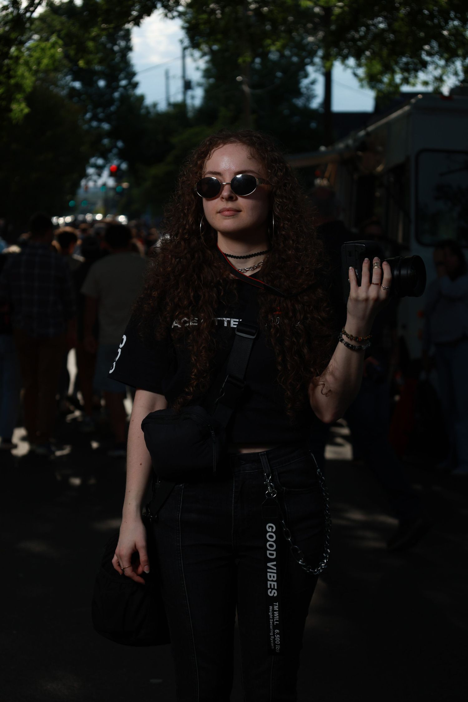 Young woman with curly hair holding a camera, wearing sunglasses and Good Vibes pants at an outdoor event.