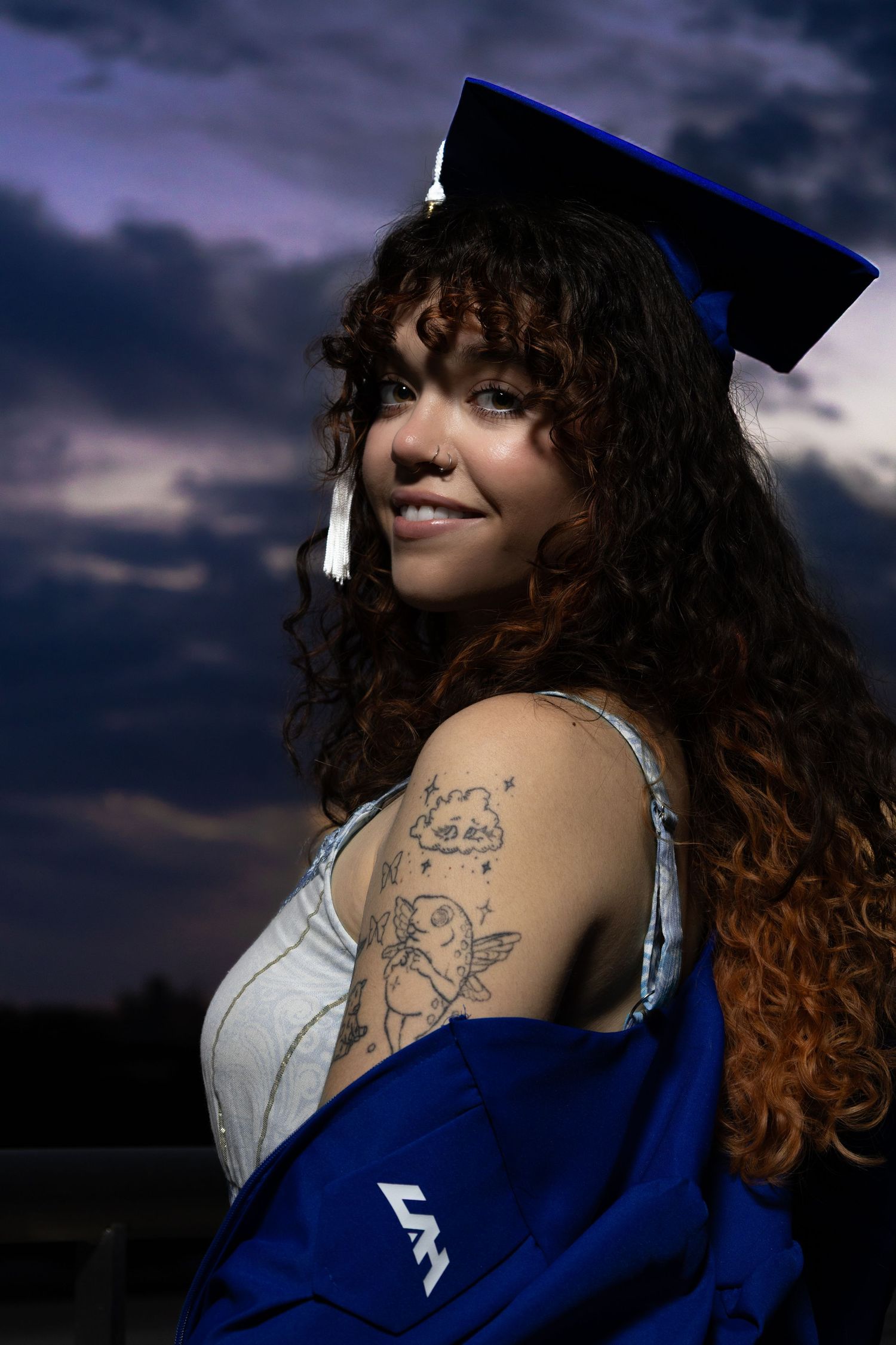 Graduate in blue cap and gown poses against dramatic sunset sky showing tattoo artwork.