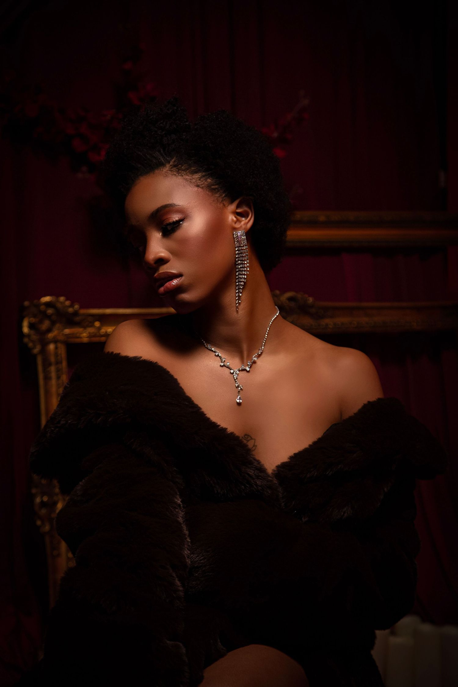 Elegant Black woman in off-shoulder fur coat with diamond necklace and earrings, posing in a dark luxurious setting.