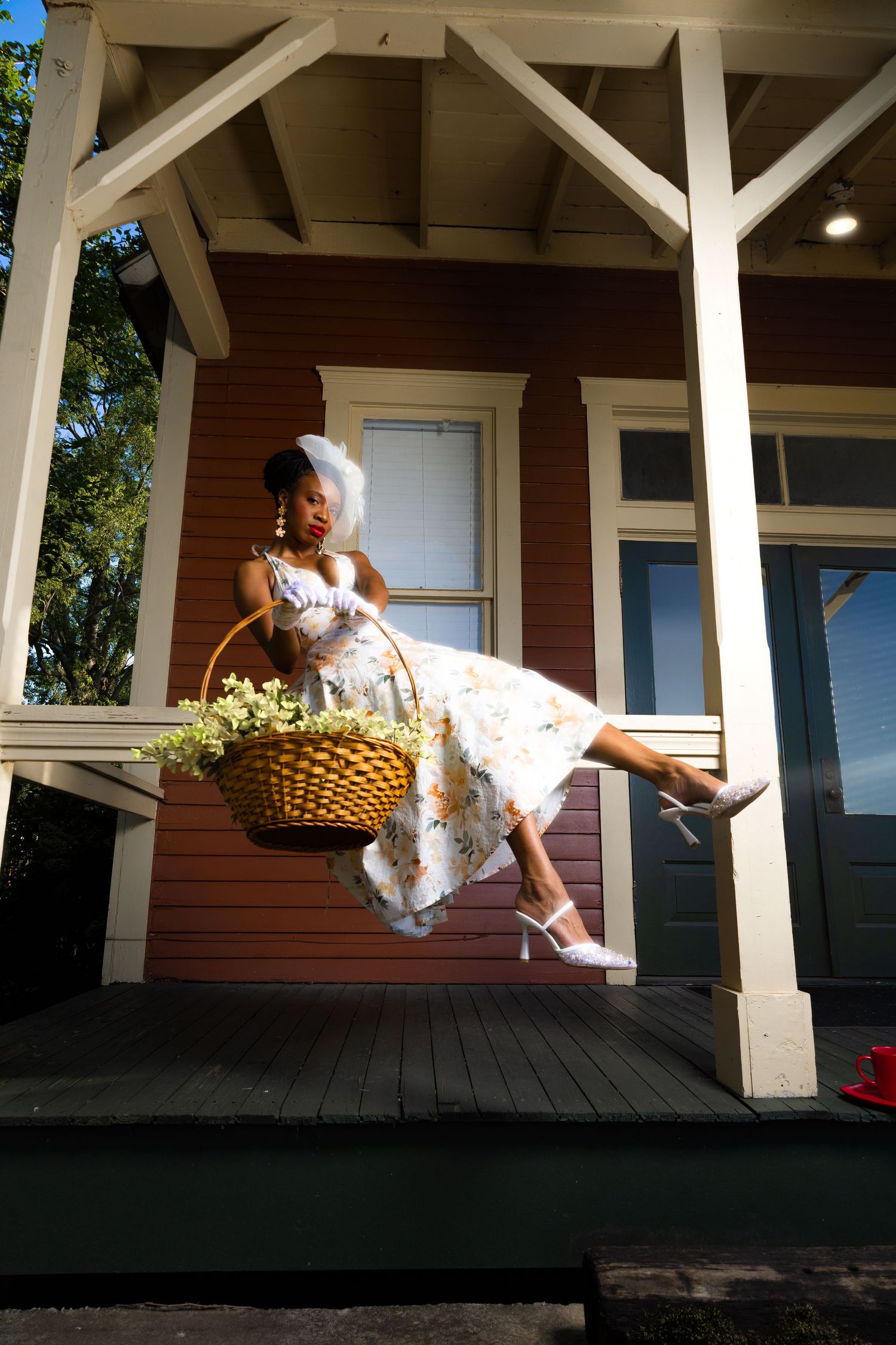 A bride in a floral dress and veil sits on porch steps holding a wicker basket filled with yellow flowers.
