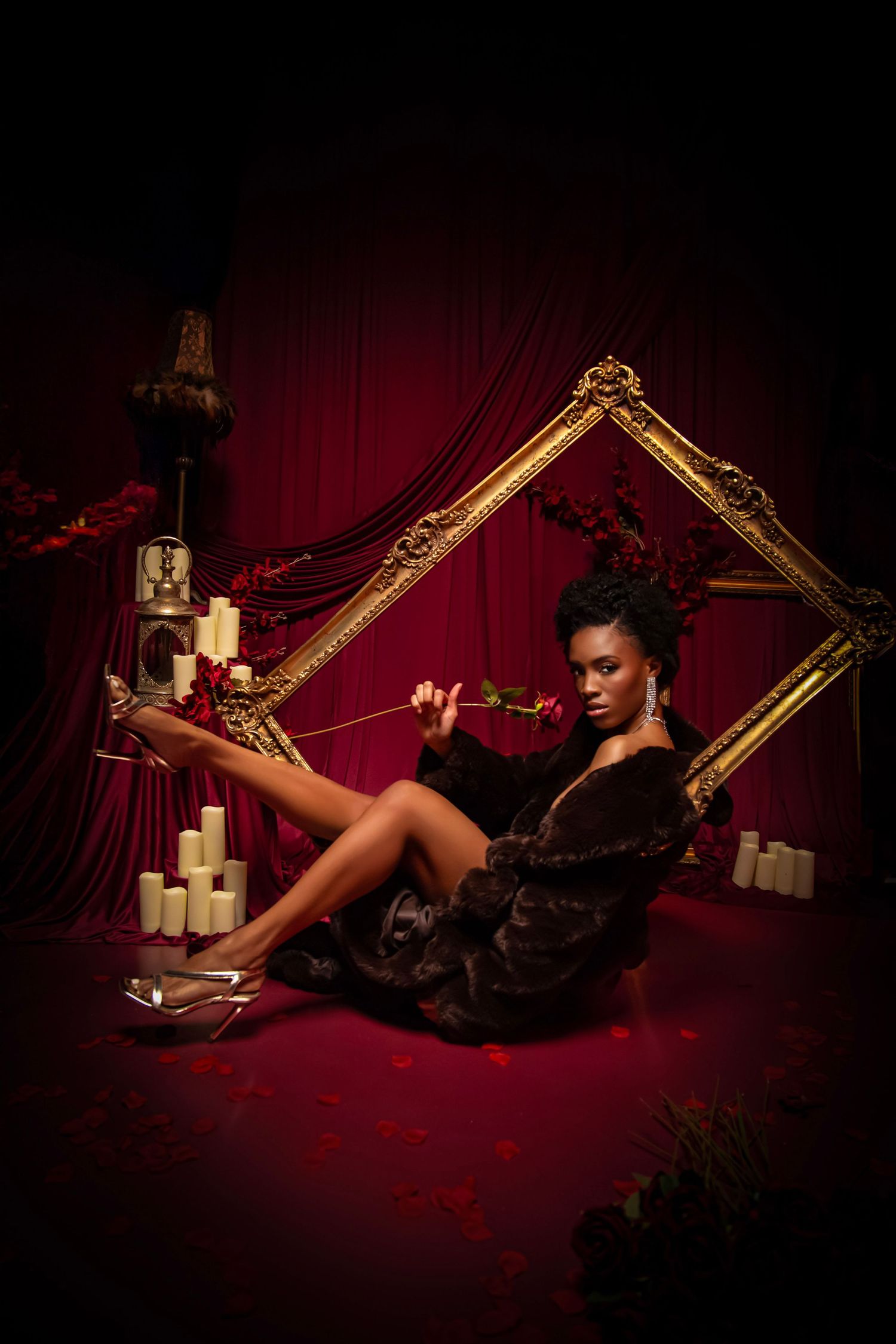 A woman in a fur coat poses with a rose beside a gold frame and candles against a dramatic red backdrop.