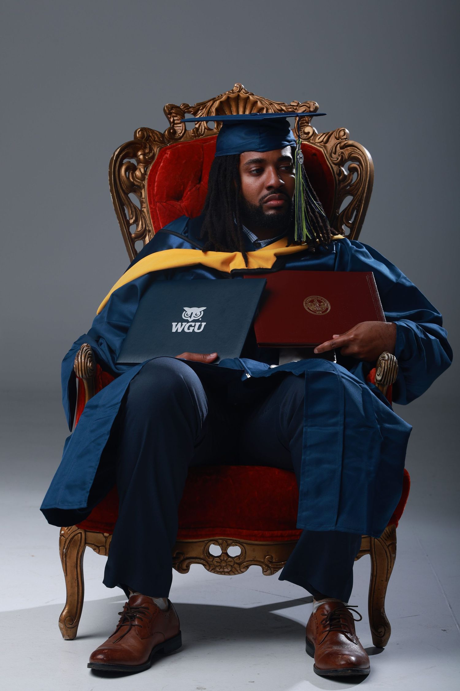 Graduate in blue cap and gown sits in ornate red throne chair holding WGU diploma and red folder.