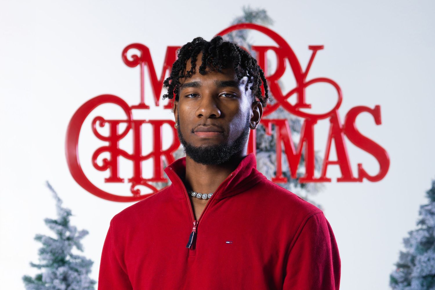 Young man in red sweater poses before a Merry Christmas sign with white trees in festive holiday photo.