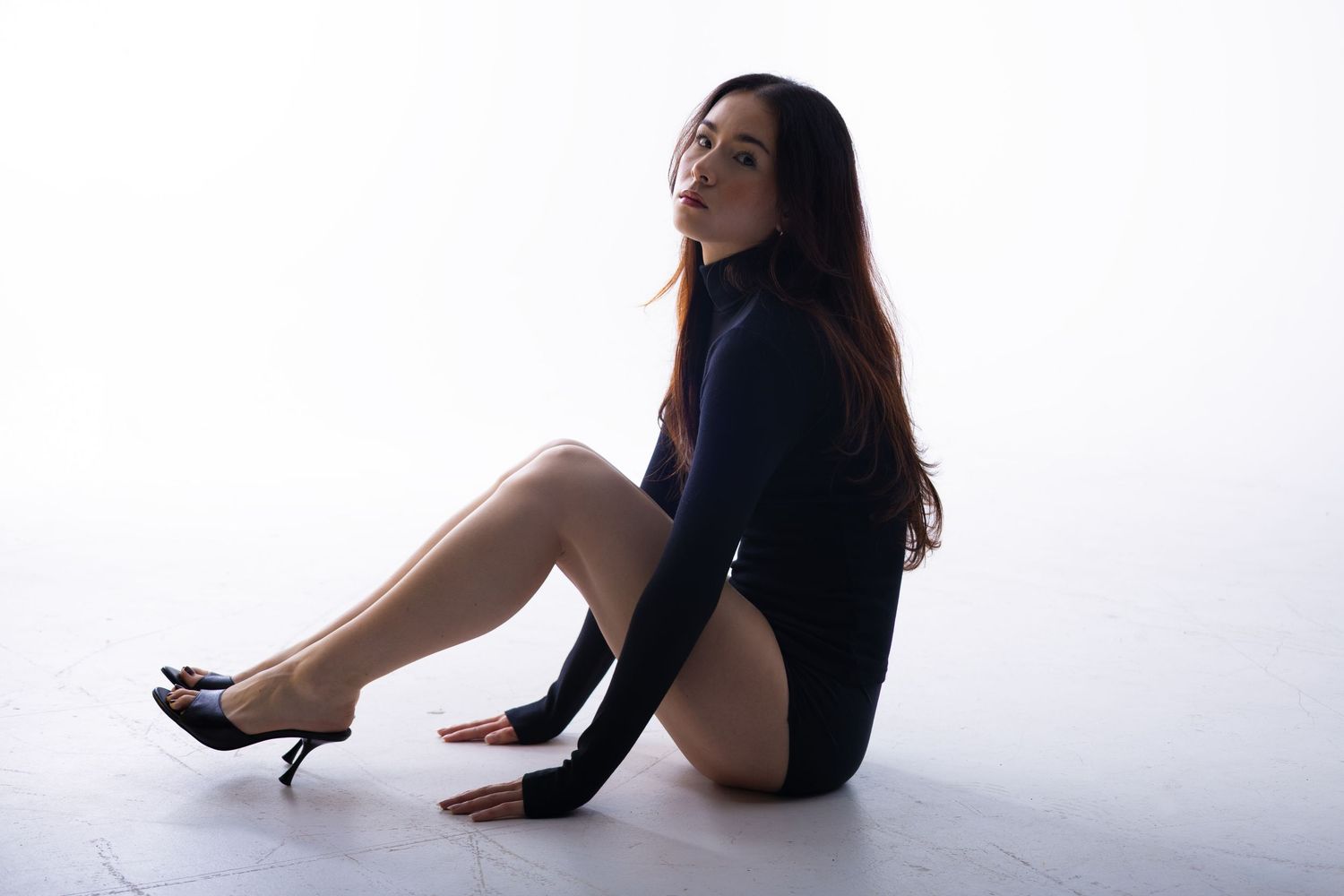 A woman in a black mini dress and heels sits gracefully on a white studio floor, posing with long dark hair.