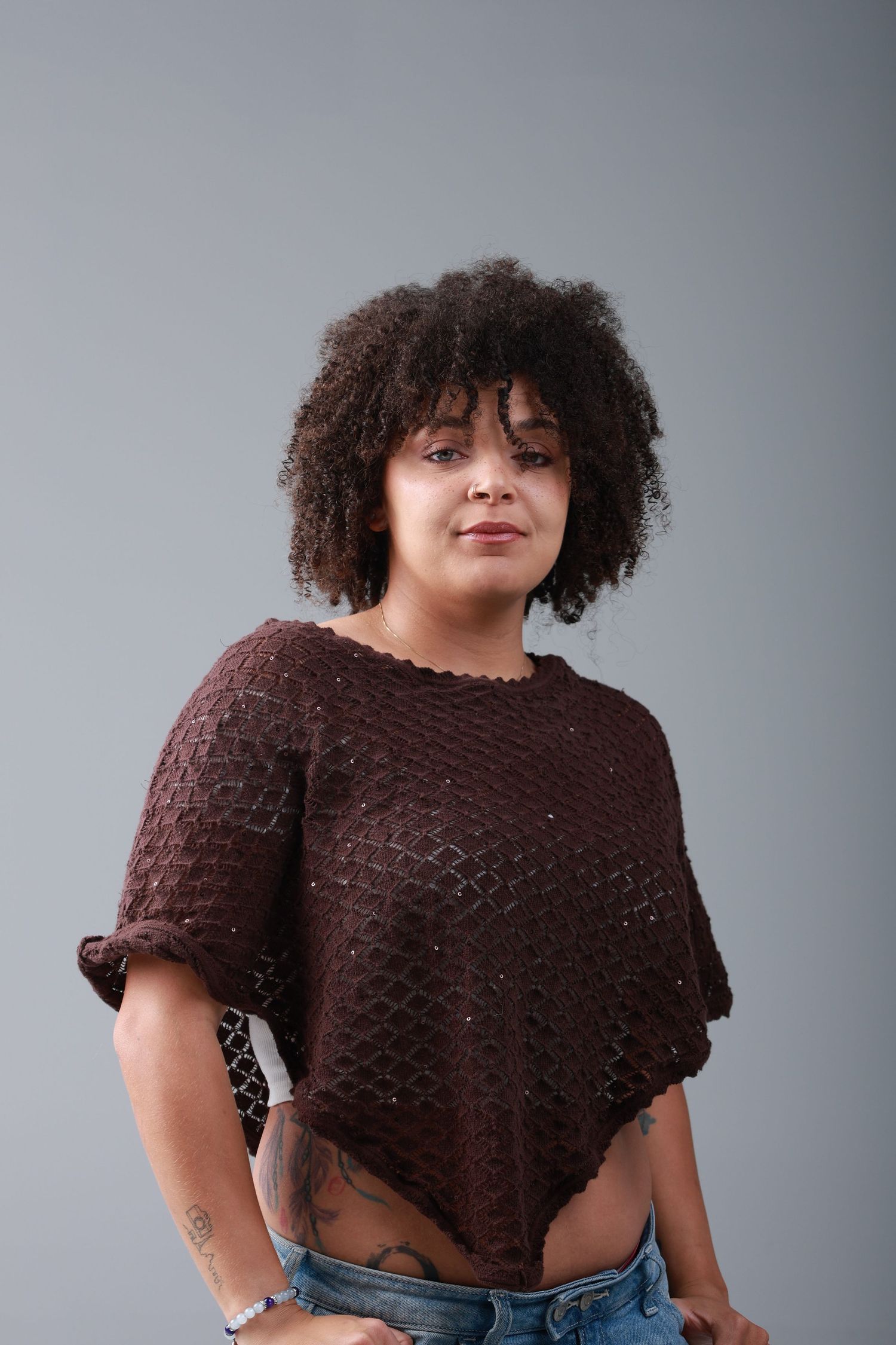A young woman with curly hair wearing a brown knit crop top and jeans poses against a gray background.