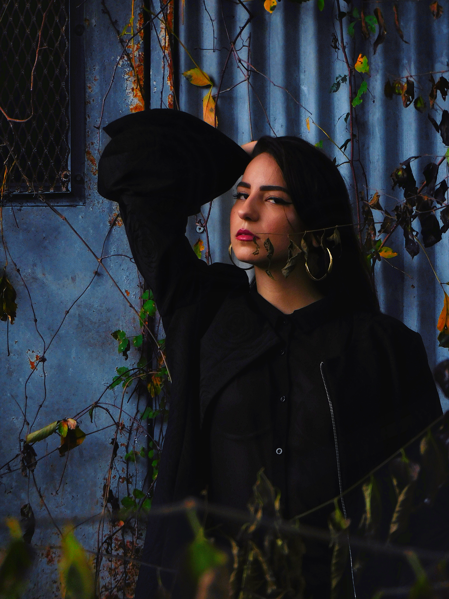 Young woman in black coat posing near rustic blue wall with autumn vines.