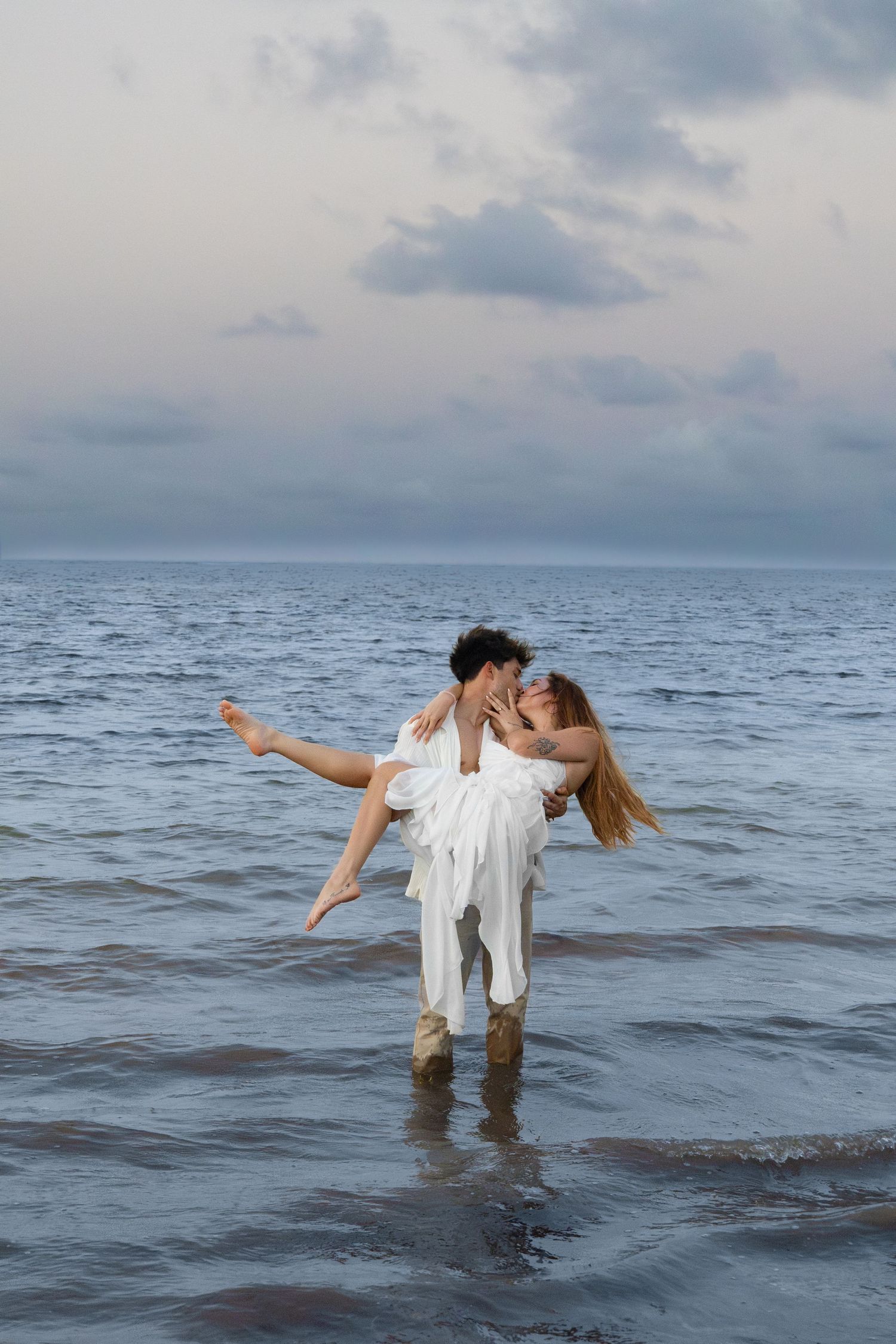Couple in white sharing a kiss while standing in ocean waves at dusk.