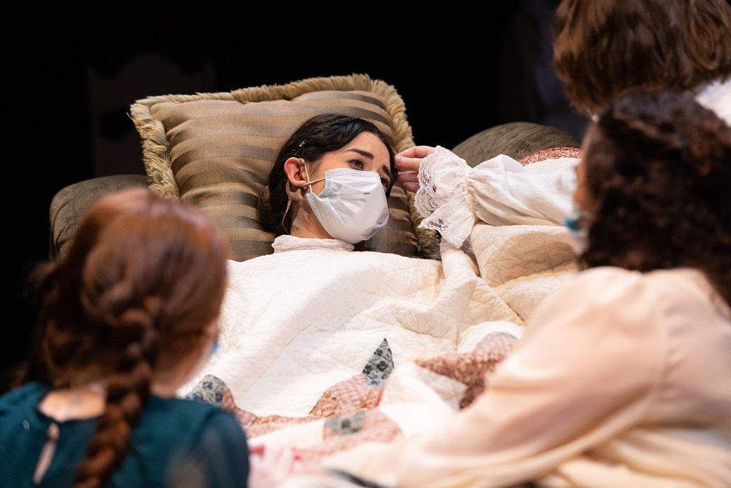 Person lying in bed wearing a white face mask surrounded by others in a theatrical stage performance.
