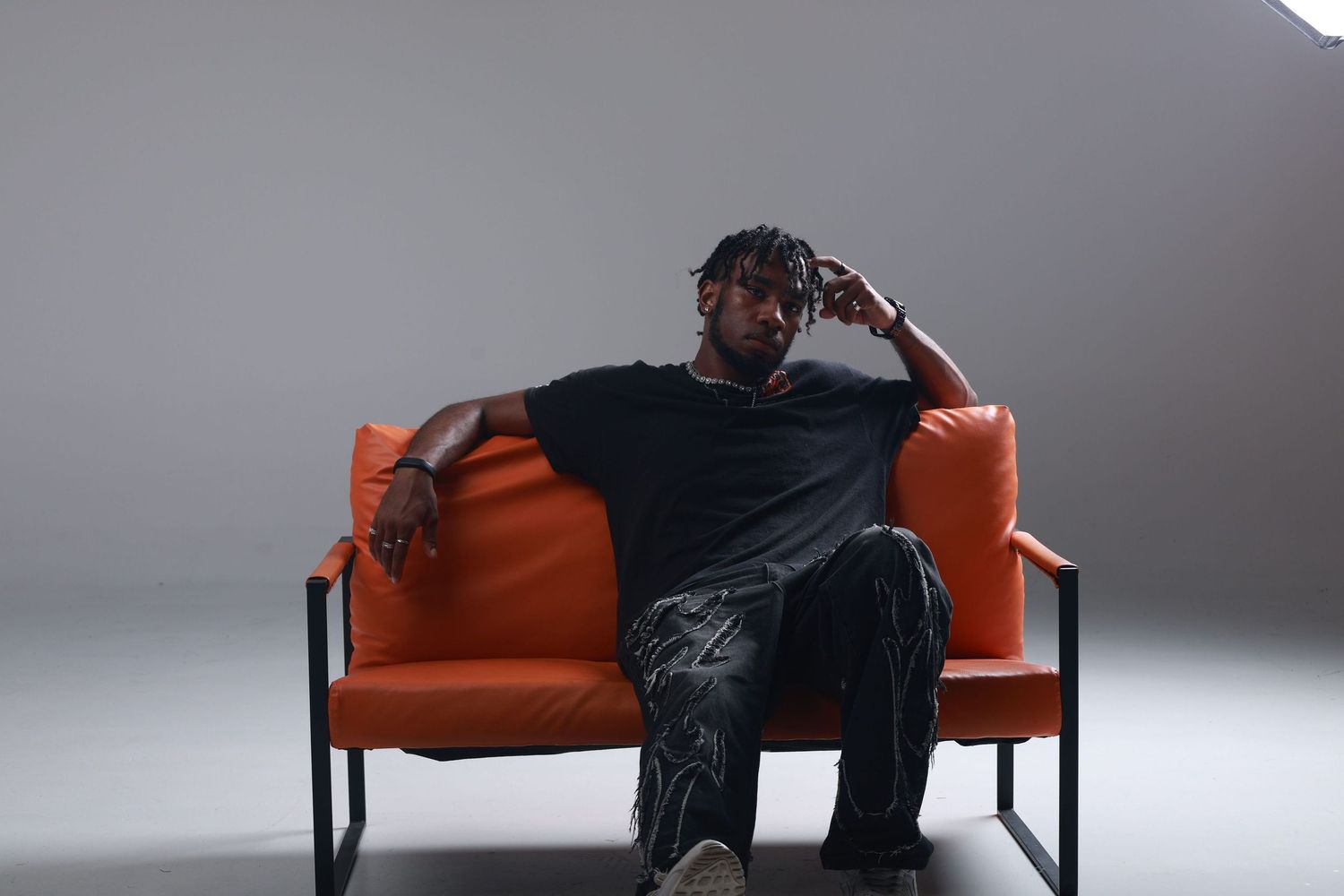 Man with dreadlocks in black t-shirt sitting relaxed on orange modern sofa against gray background.