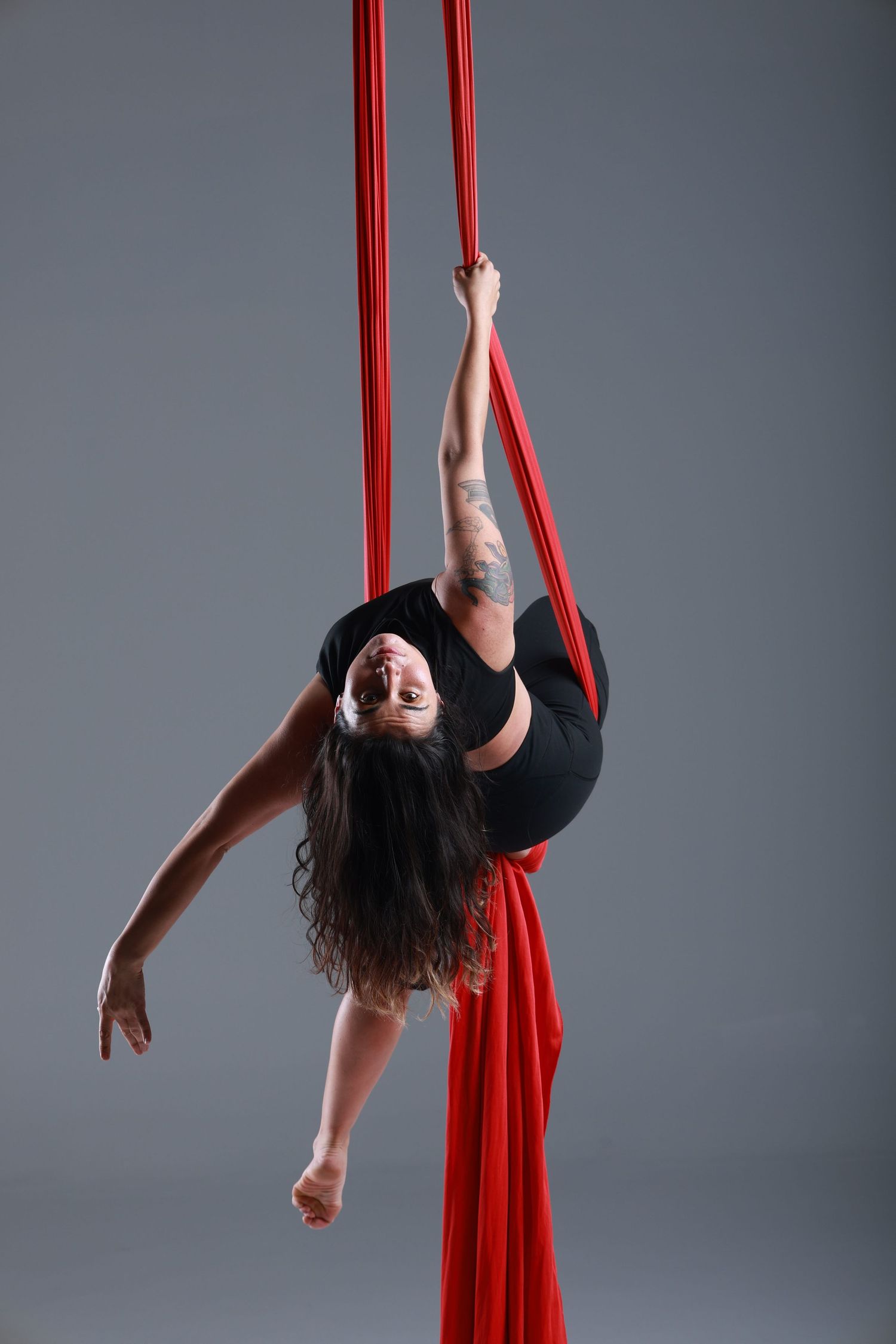 Aerial silk performer in black outfit gracefully backbends while holding red silks against gray background.