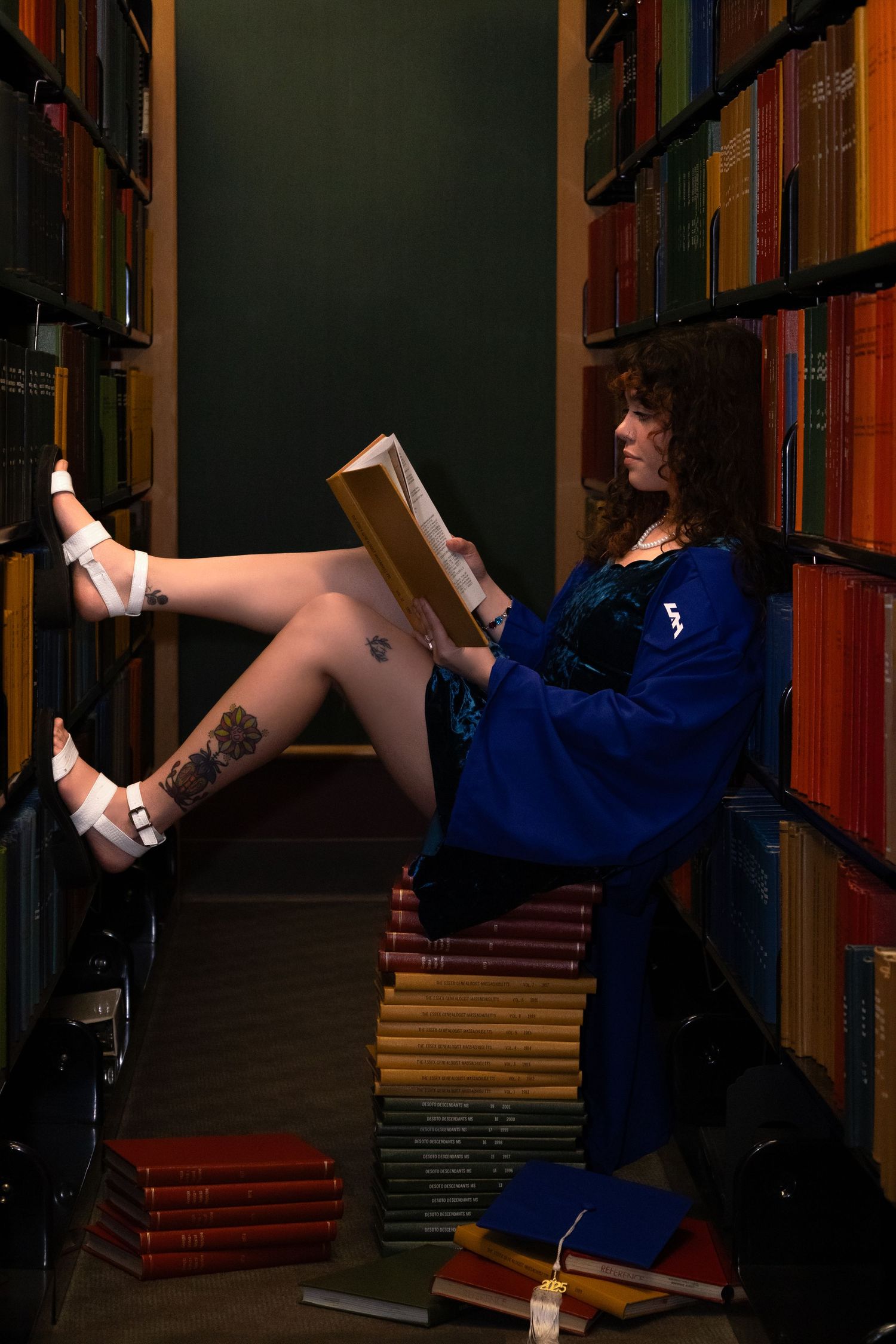 Person nestled between library shelves in a blue dress reading a book while reclining on stacks of vintage volumes.