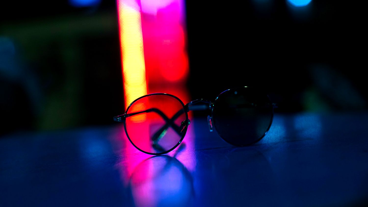 Round sunglasses resting on a reflective surface with vibrant pink, red, and blue neon lights in the background.