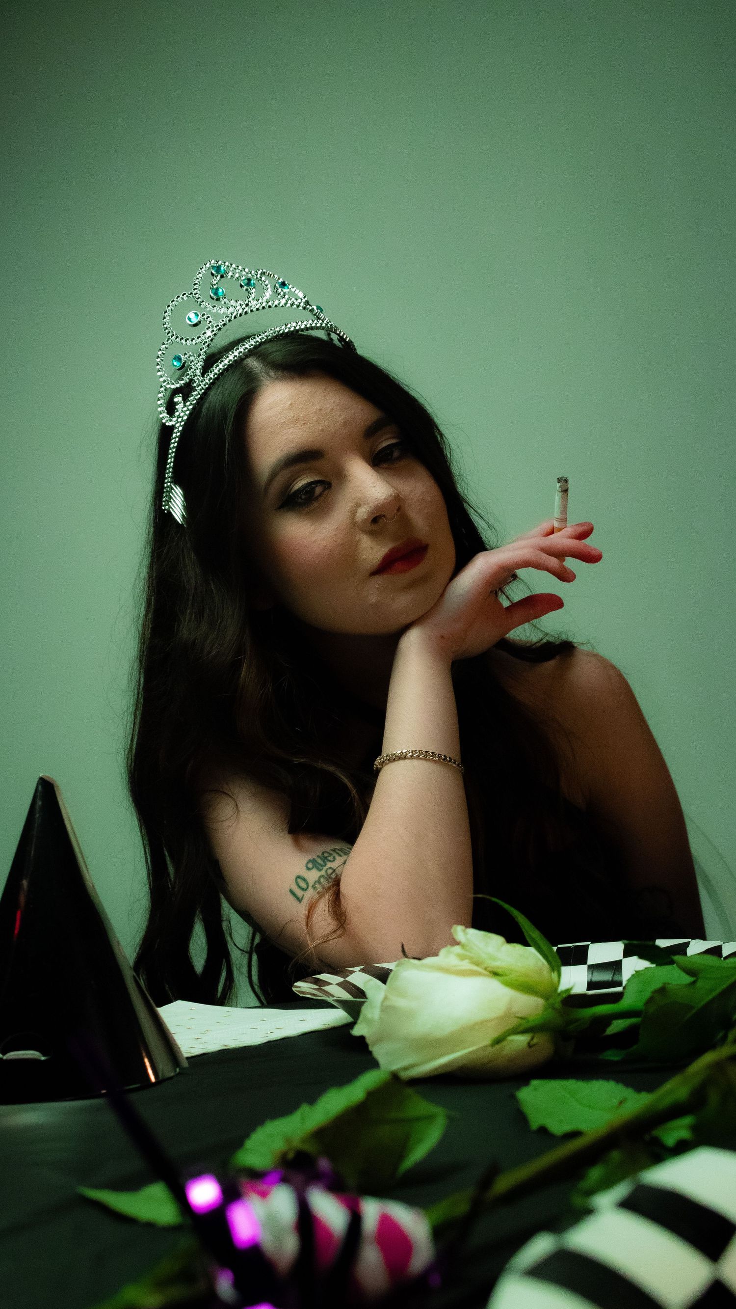 Young woman wearing silver tiara and holding cigarette sitting at table with white rose in moody green light.