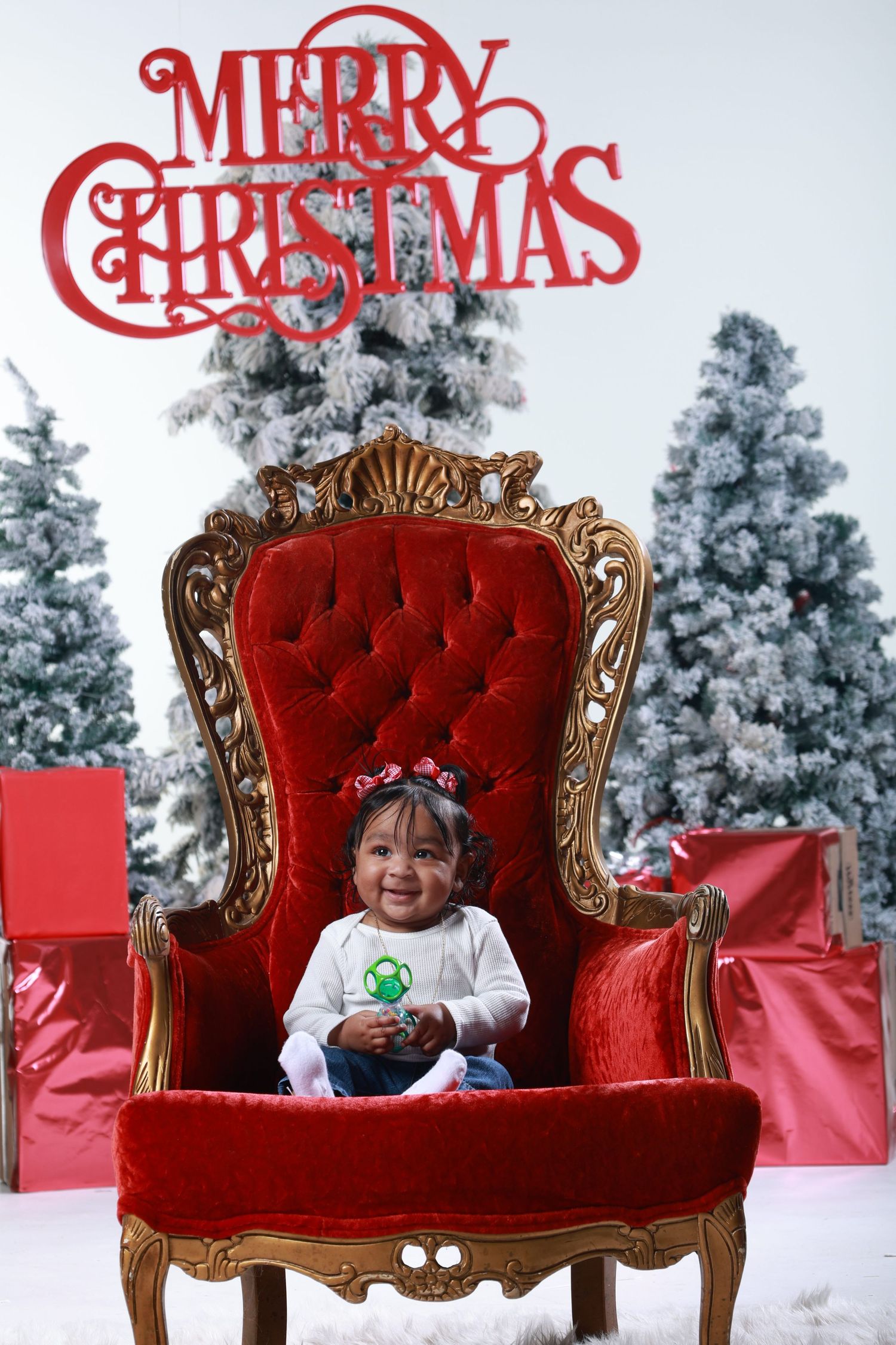 A smiling baby girl sits in a grand red velvet throne chair with a Merry Christmas sign and snowy trees behind her.