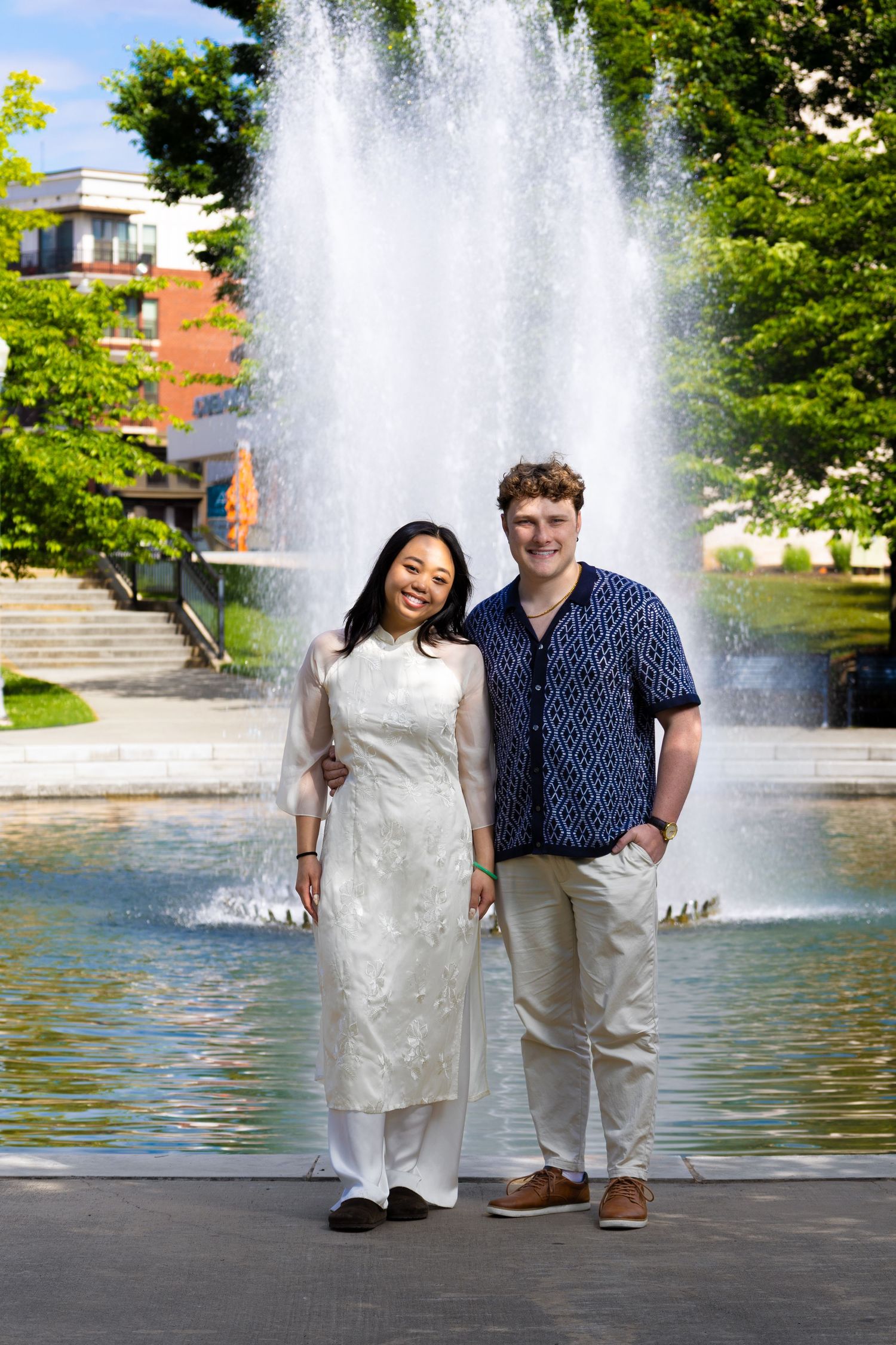 Two people standing together in front of a large water fountain on a sunny day in a park setting.