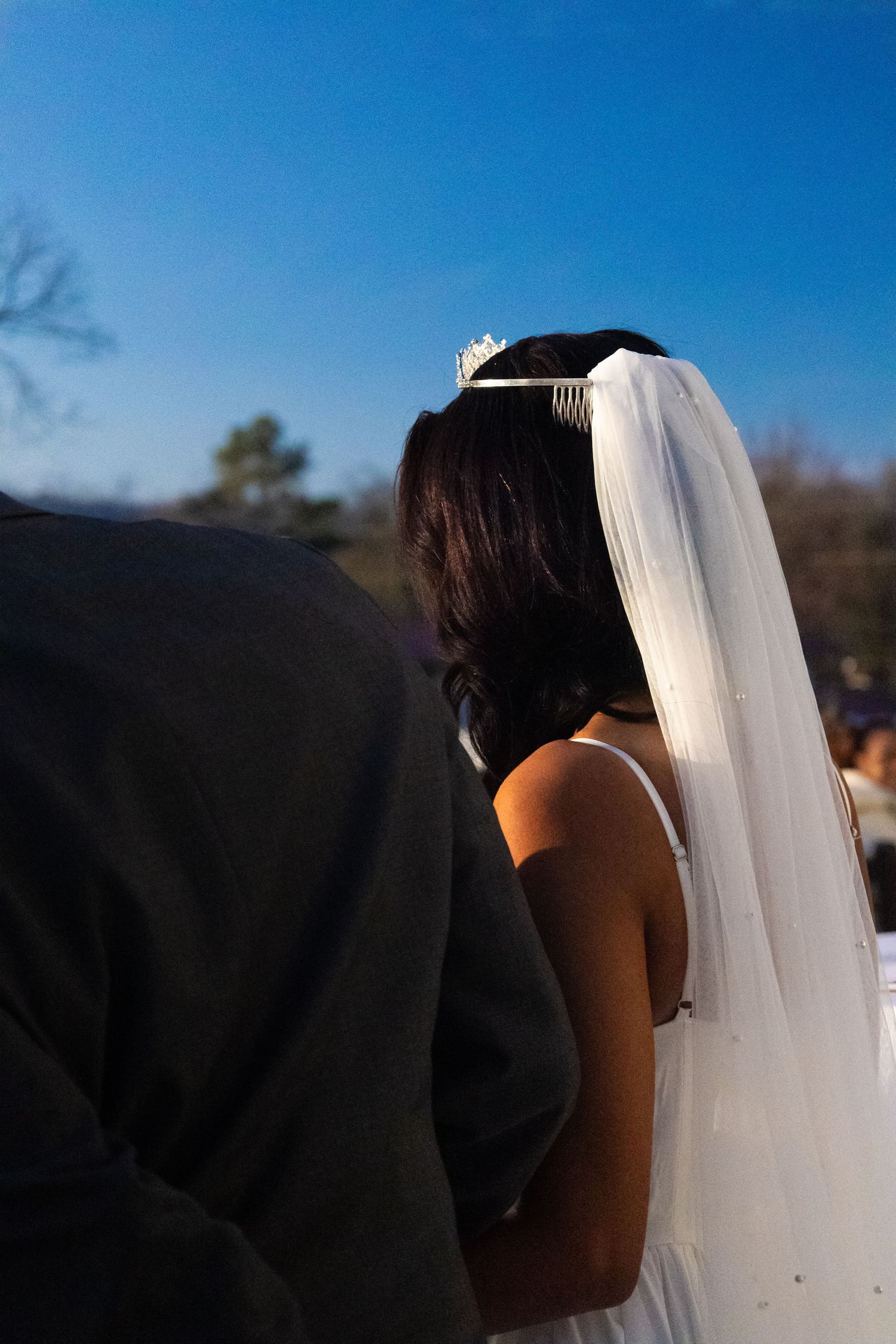 Bride wearing white veil and tiara stands beside groom outdoors under clear blue sky at wedding ceremony.