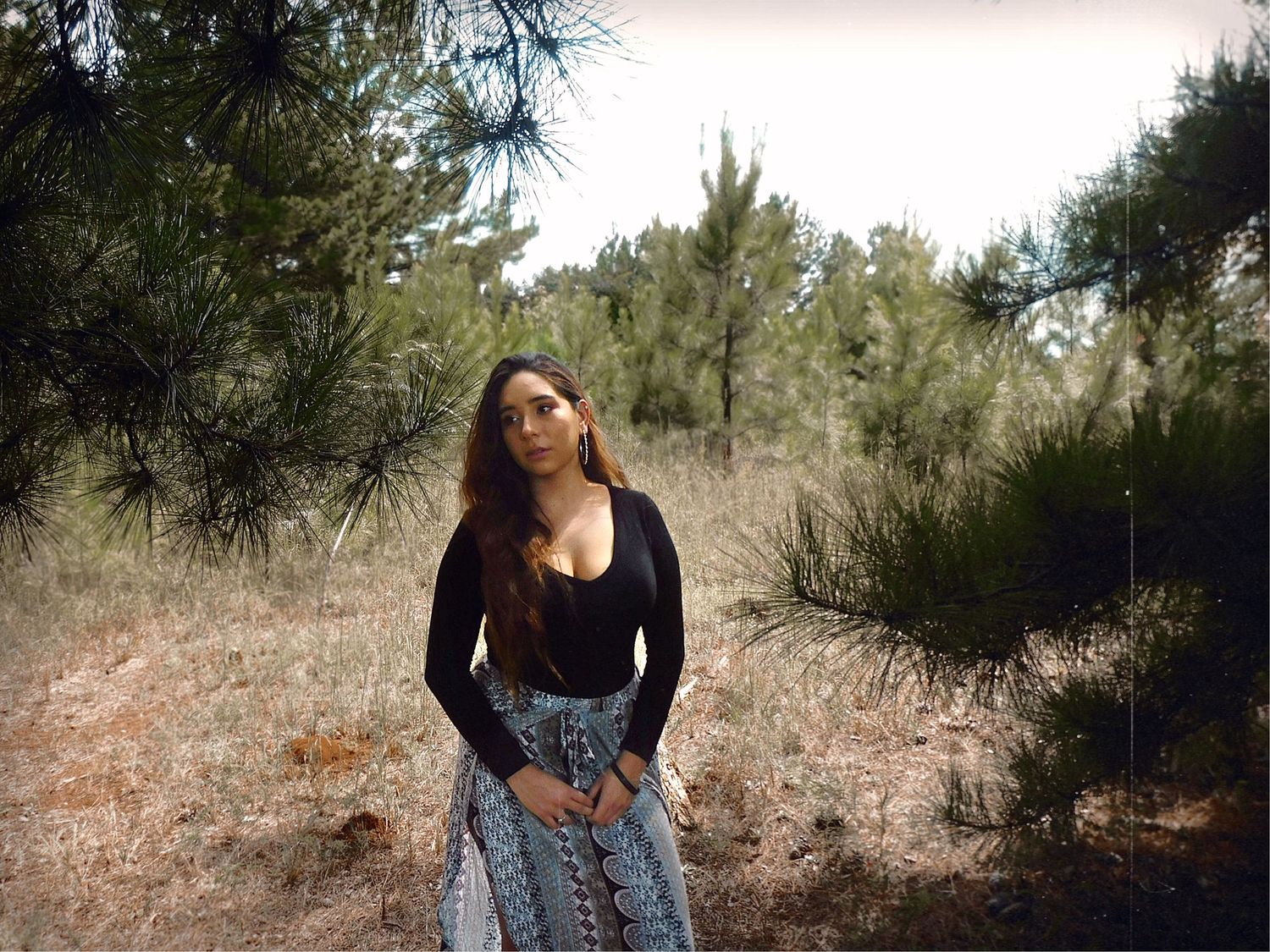 A young woman in a black top and patterned skirt stands among pine trees in a natural forest setting.
