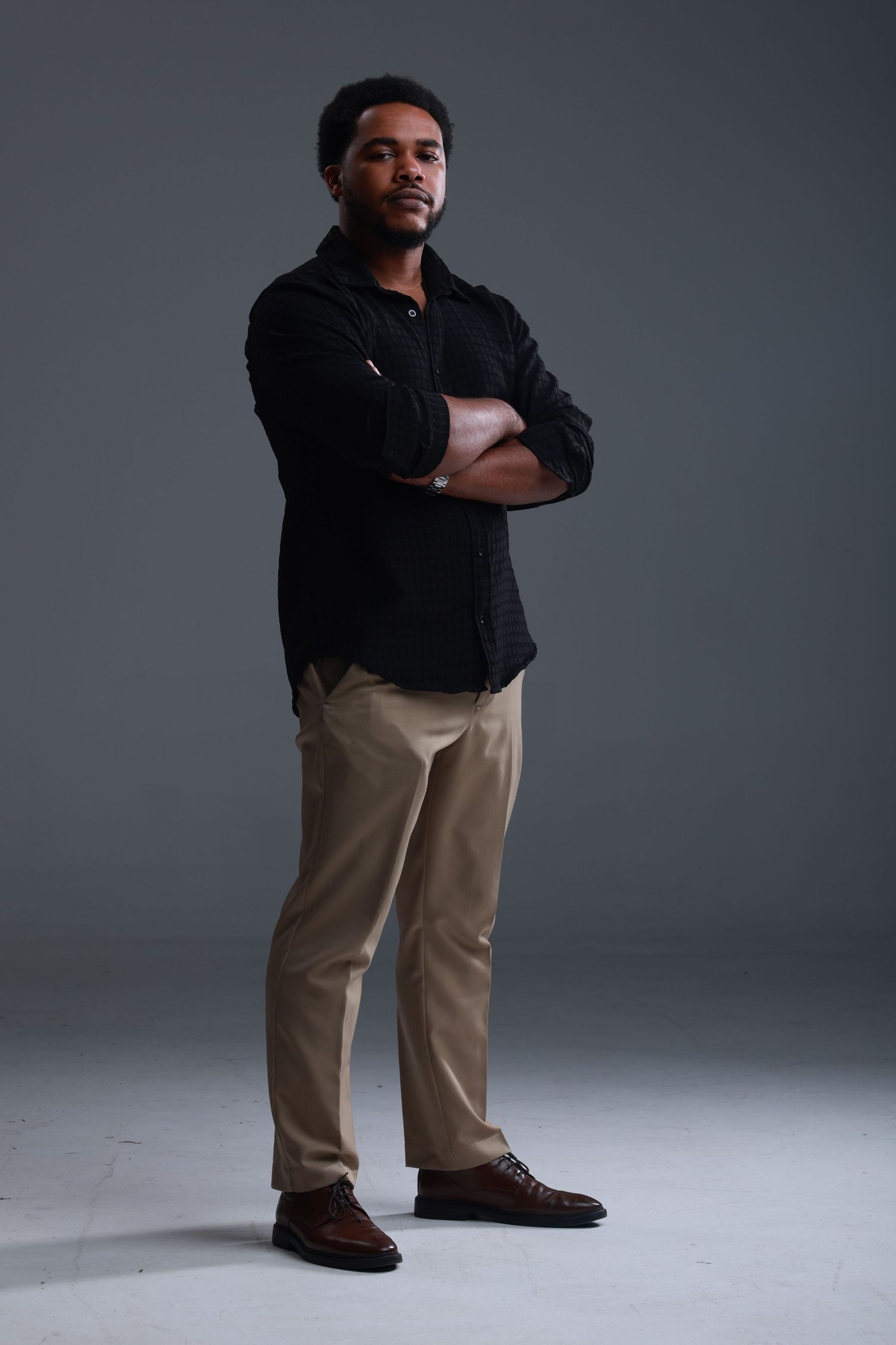 A confident young Black man in a black shirt and khaki pants stands with arms crossed against a gray studio background.