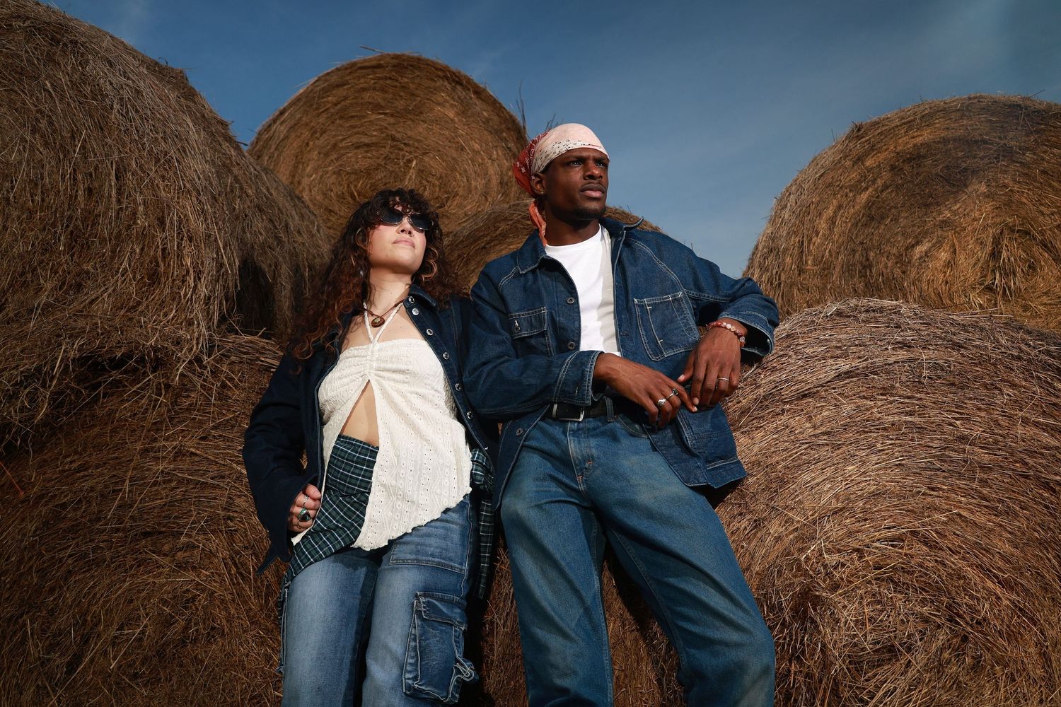 Two models in denim outfits pose against large hay bales under a blue sky in a rustic outdoor setting.