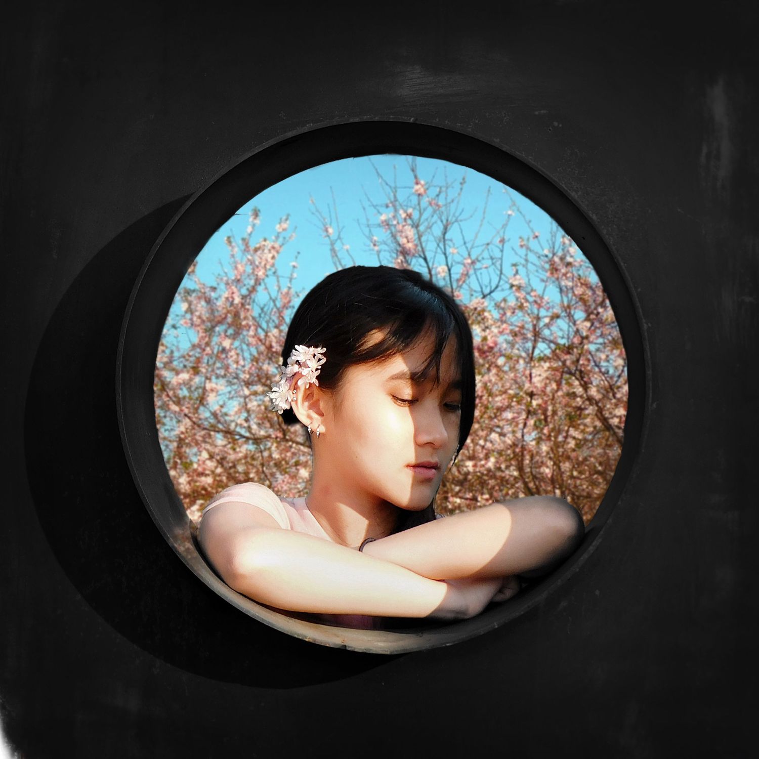 A young woman with a flower in her hair poses in a circular window framed by blooming cherry blossom trees.