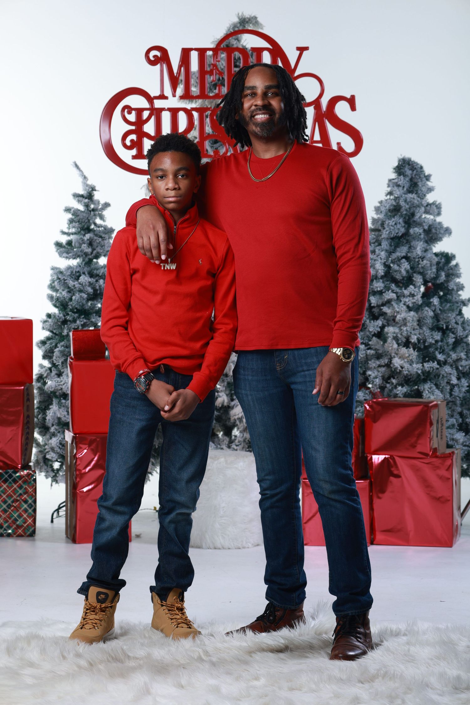 Father and son in matching red sweaters and jeans pose for a festive Christmas photo with decorated trees and gifts.