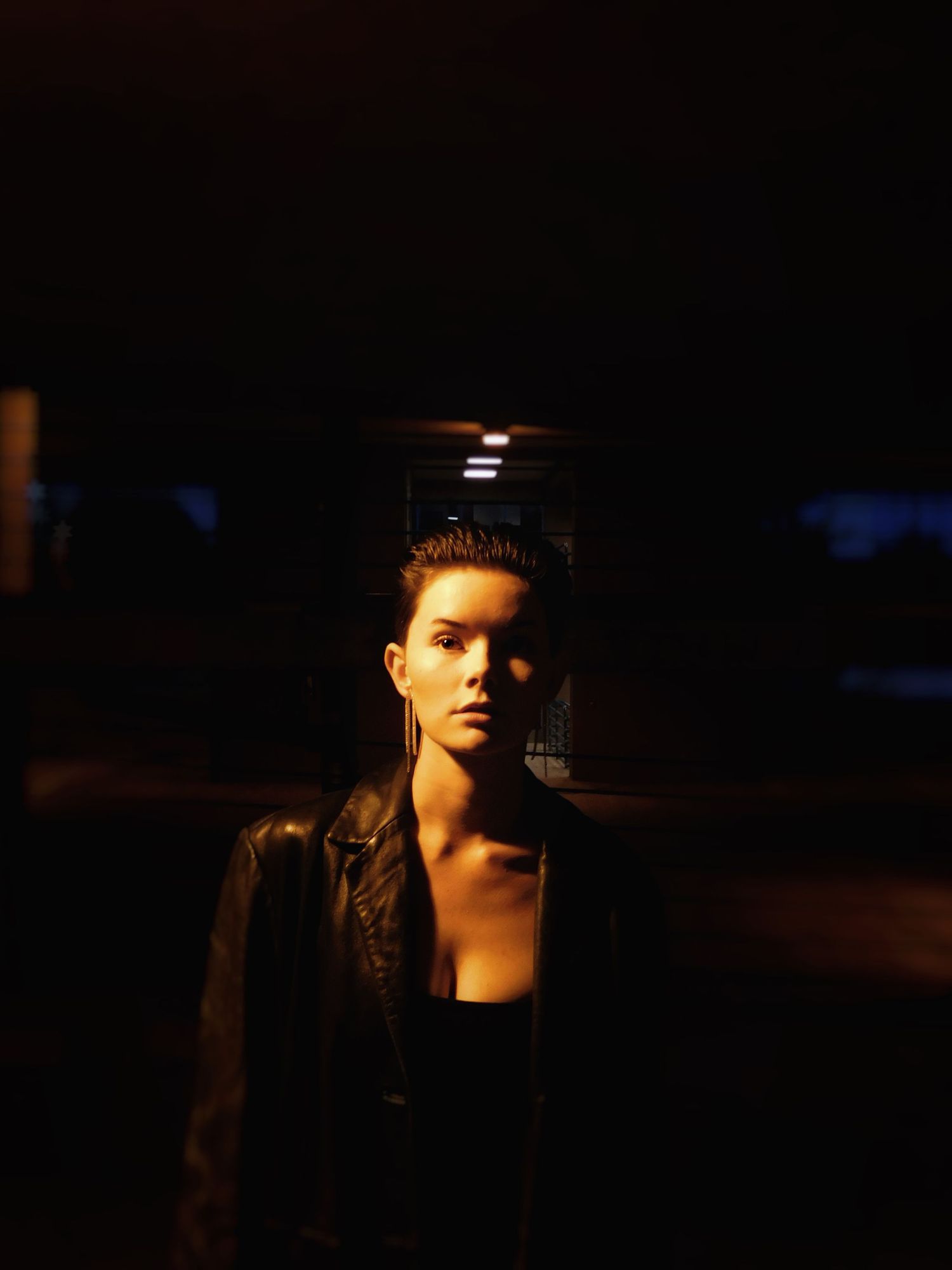 A woman in a leather jacket stands in a dark urban setting illuminated by warm artificial lighting.