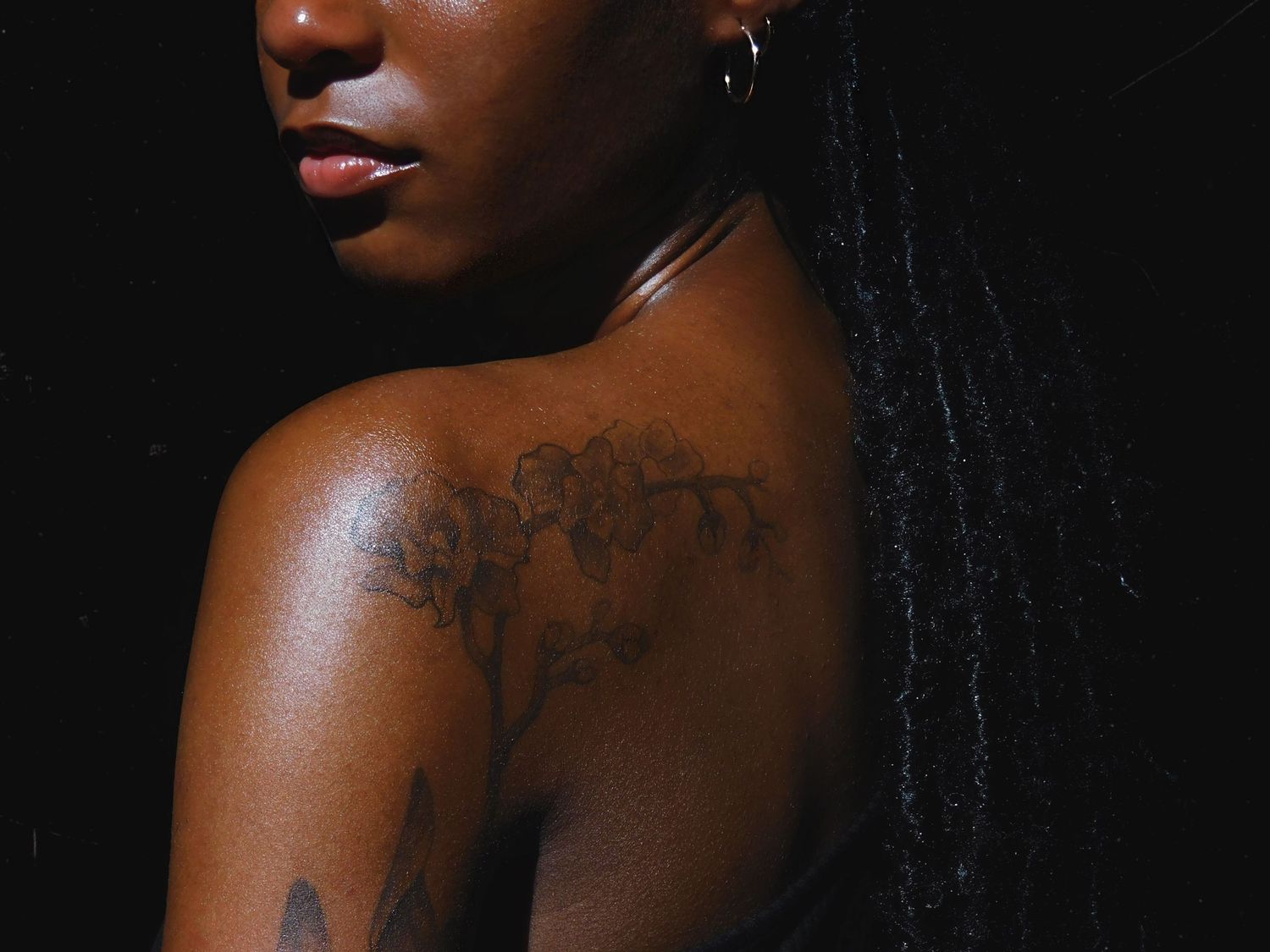 A young Black woman with braids showcasing floral tattoos on her shoulder and back against a dark background.