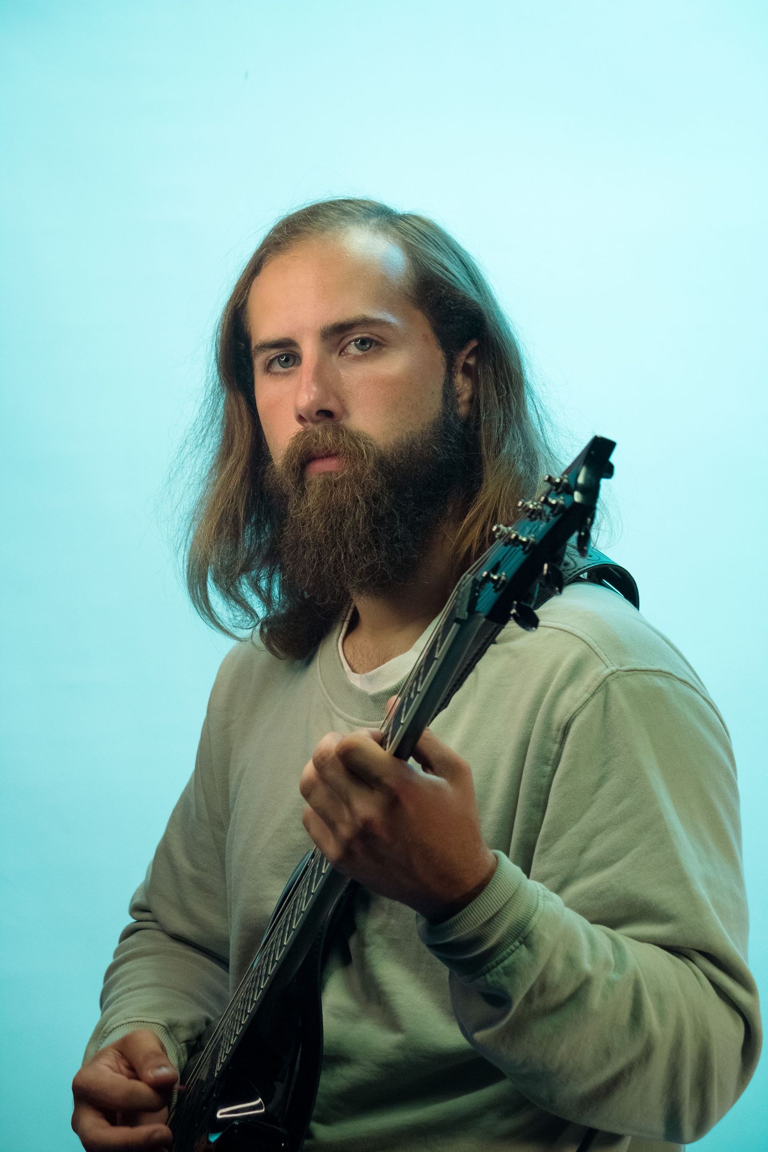 Bearded man with long hair playing black electric guitar against teal background.