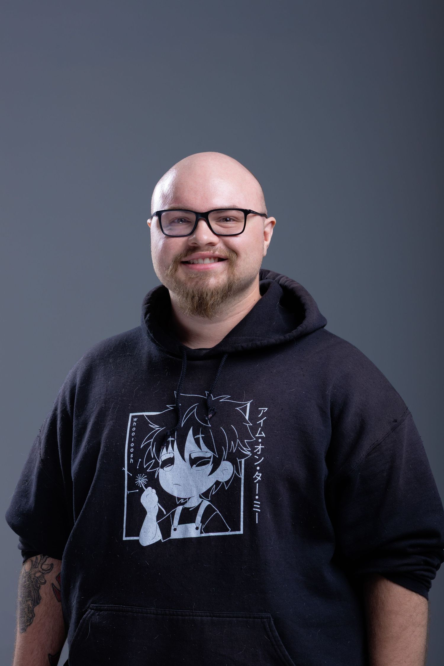 Bald man with glasses and beard wearing black anime hoodie smiling against gray background.
