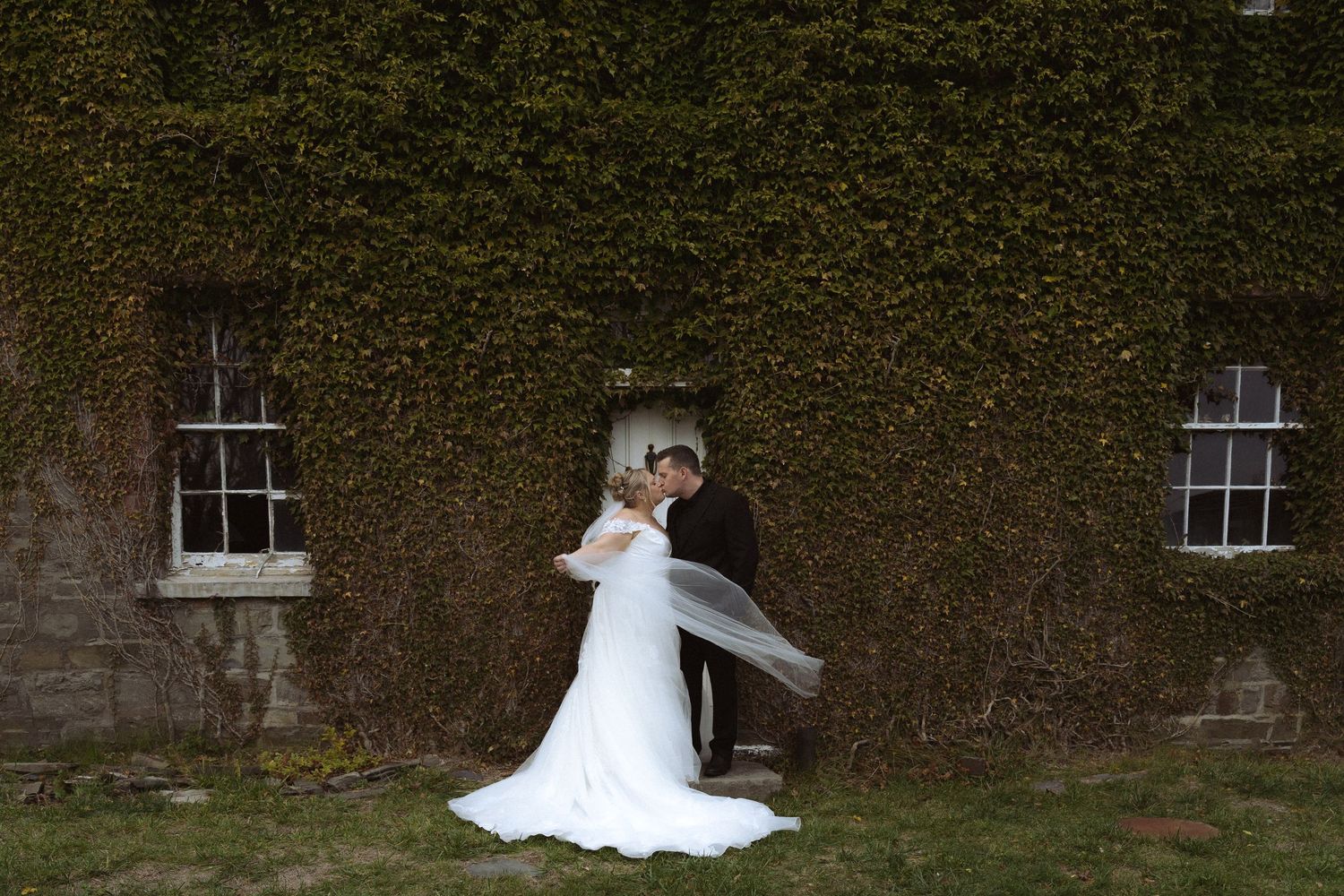A couple in wedding attire dances together against an ivy-covered stone wall with two windows.