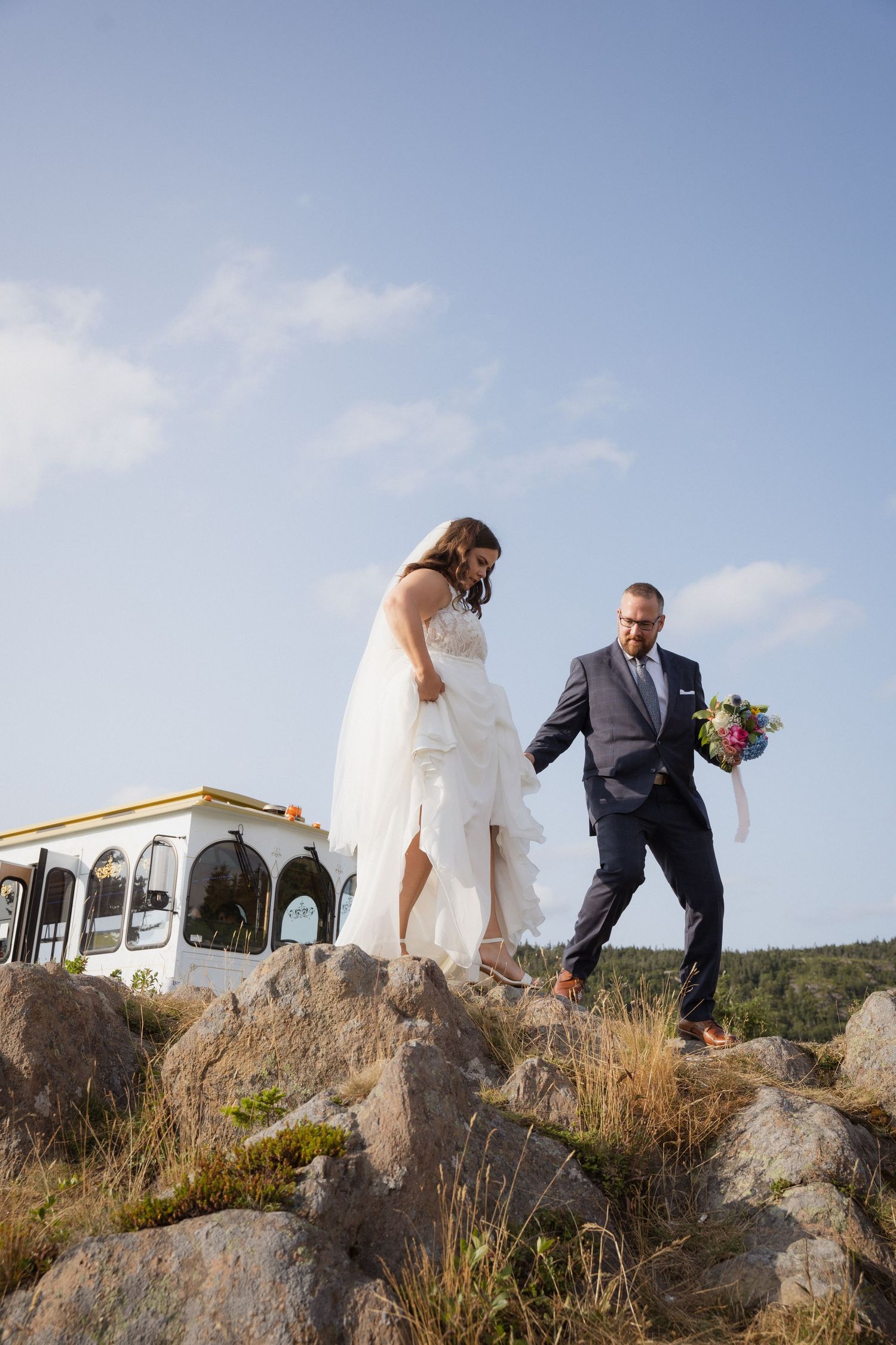 A wedding couple poses on rocky terrain with a white building in the background.