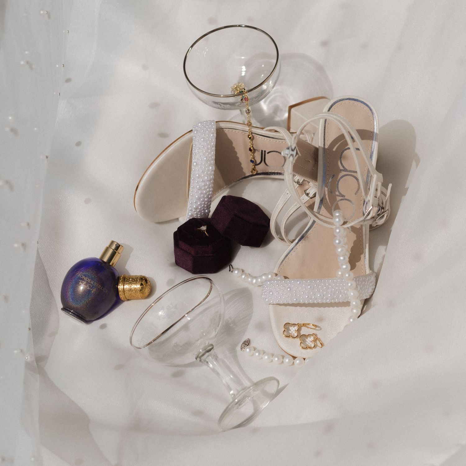 Elegant white strappy sandals arranged with wine glasses and purple light bulbs on a white textured surface.