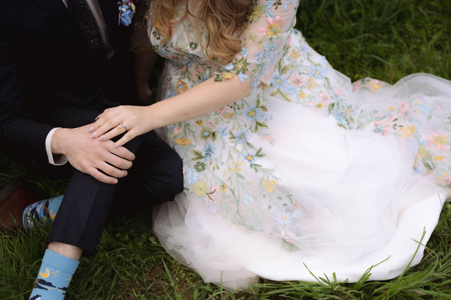 Wedding couple in white dress and dark suit embracing on grass with floral details.