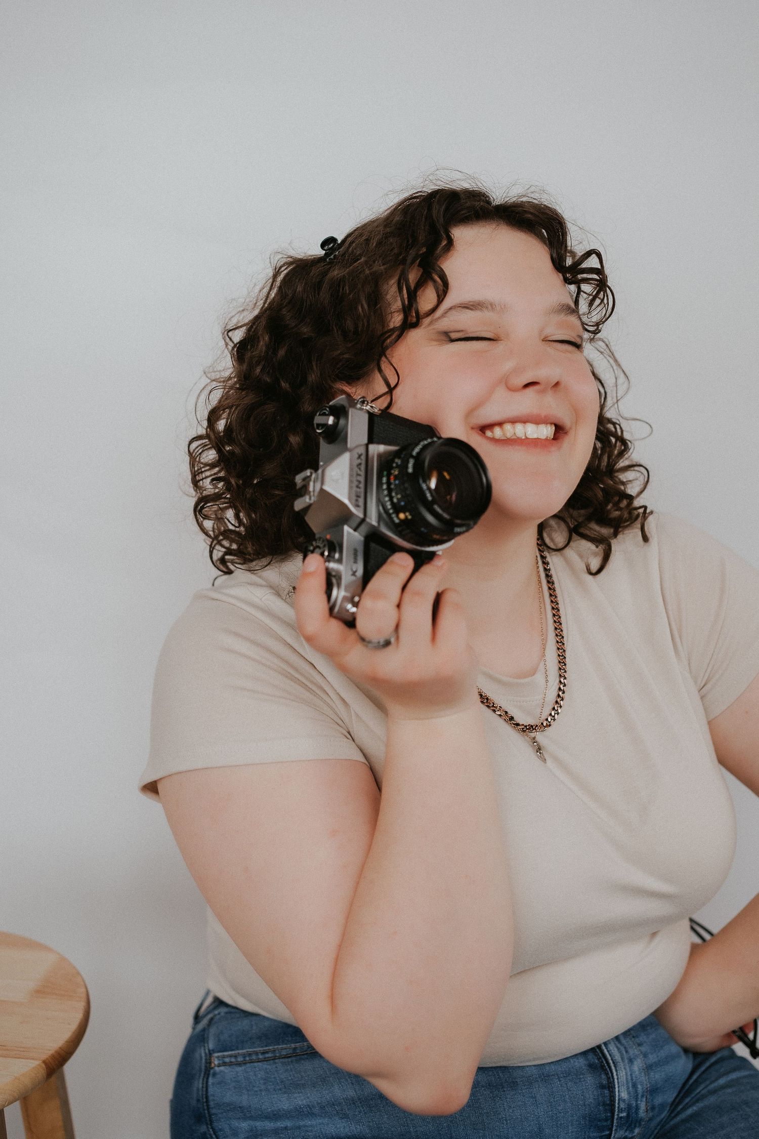 A person with curly hair laughing while holding a vintage camera against a grey background.