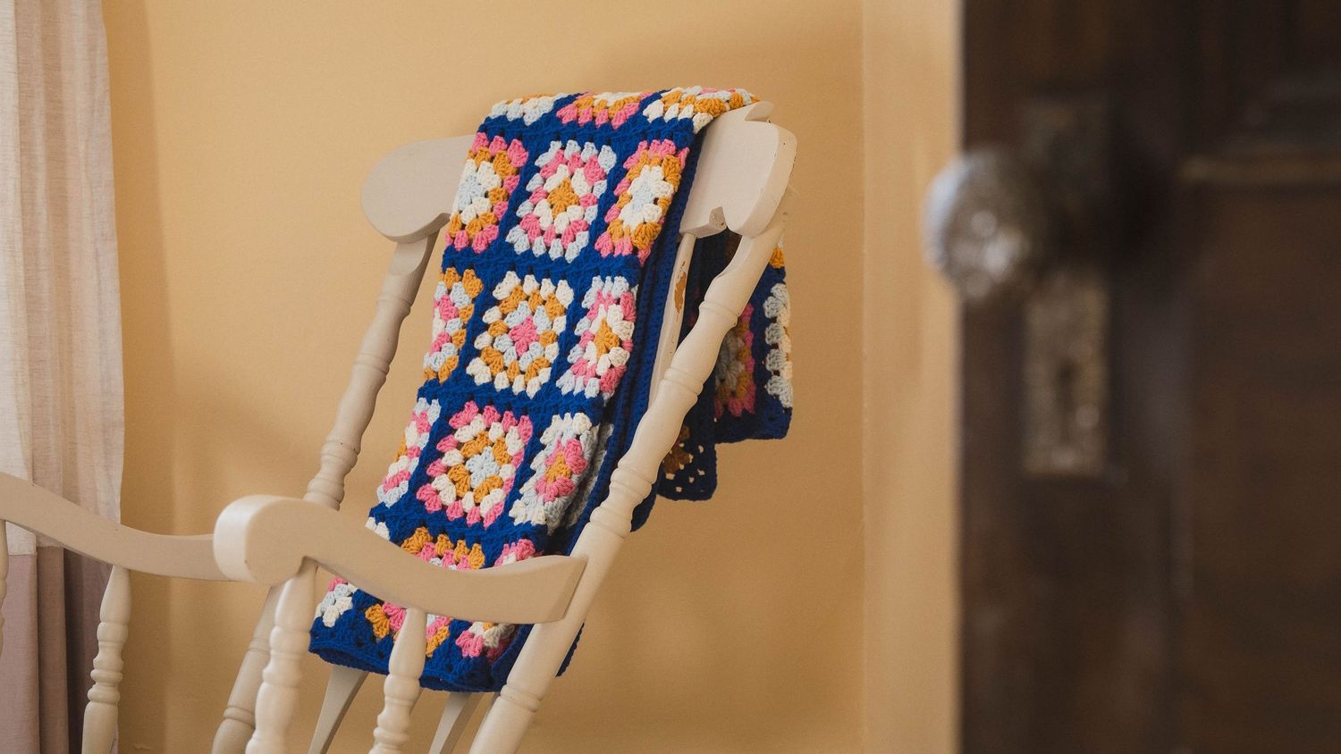 Colorful crocheted granny square blanket draped over a white wooden rocking chair near a door.