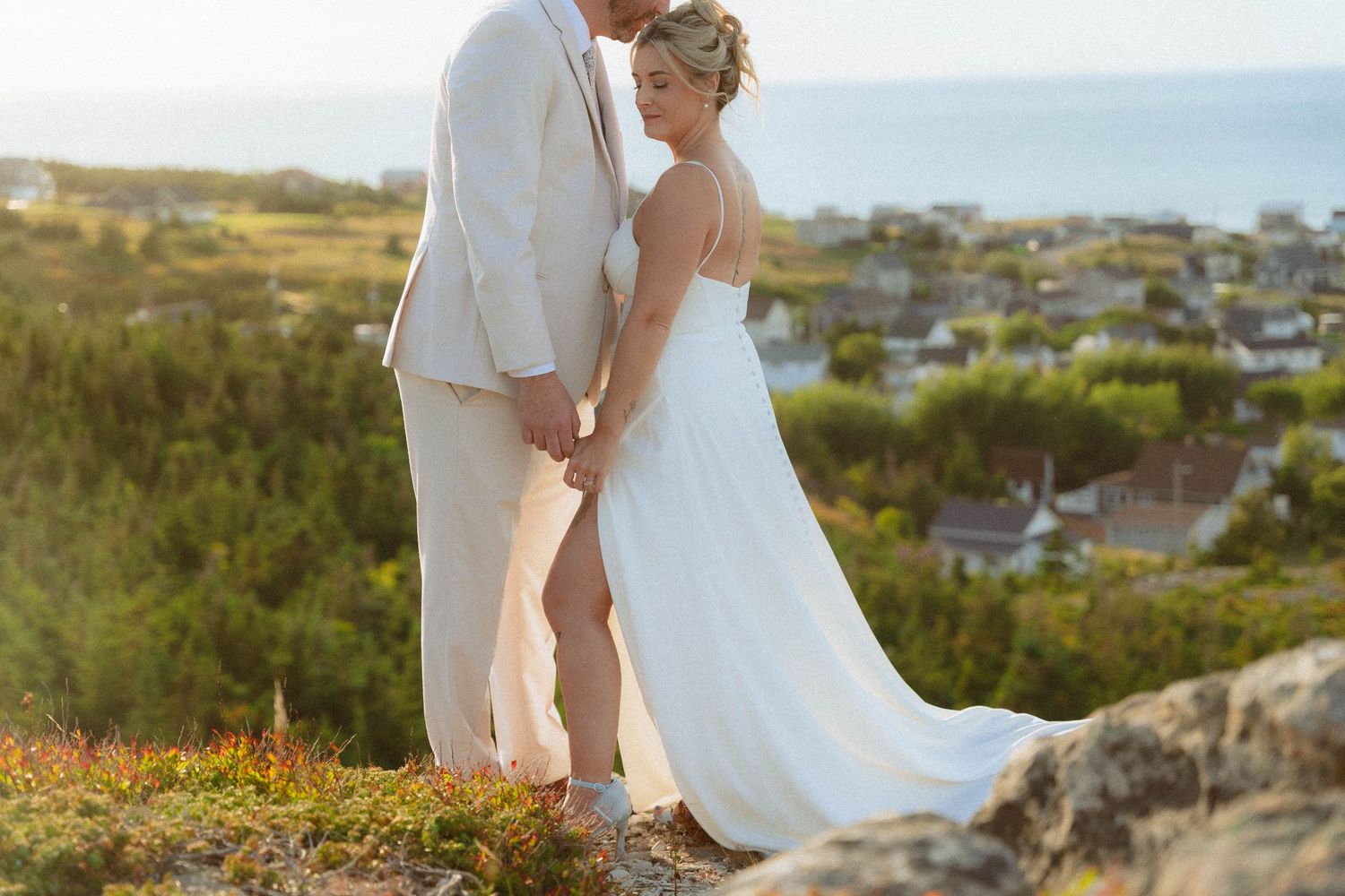 A couple in wedding attire stands on a hilltop overlooking a coastal town during golden hour.