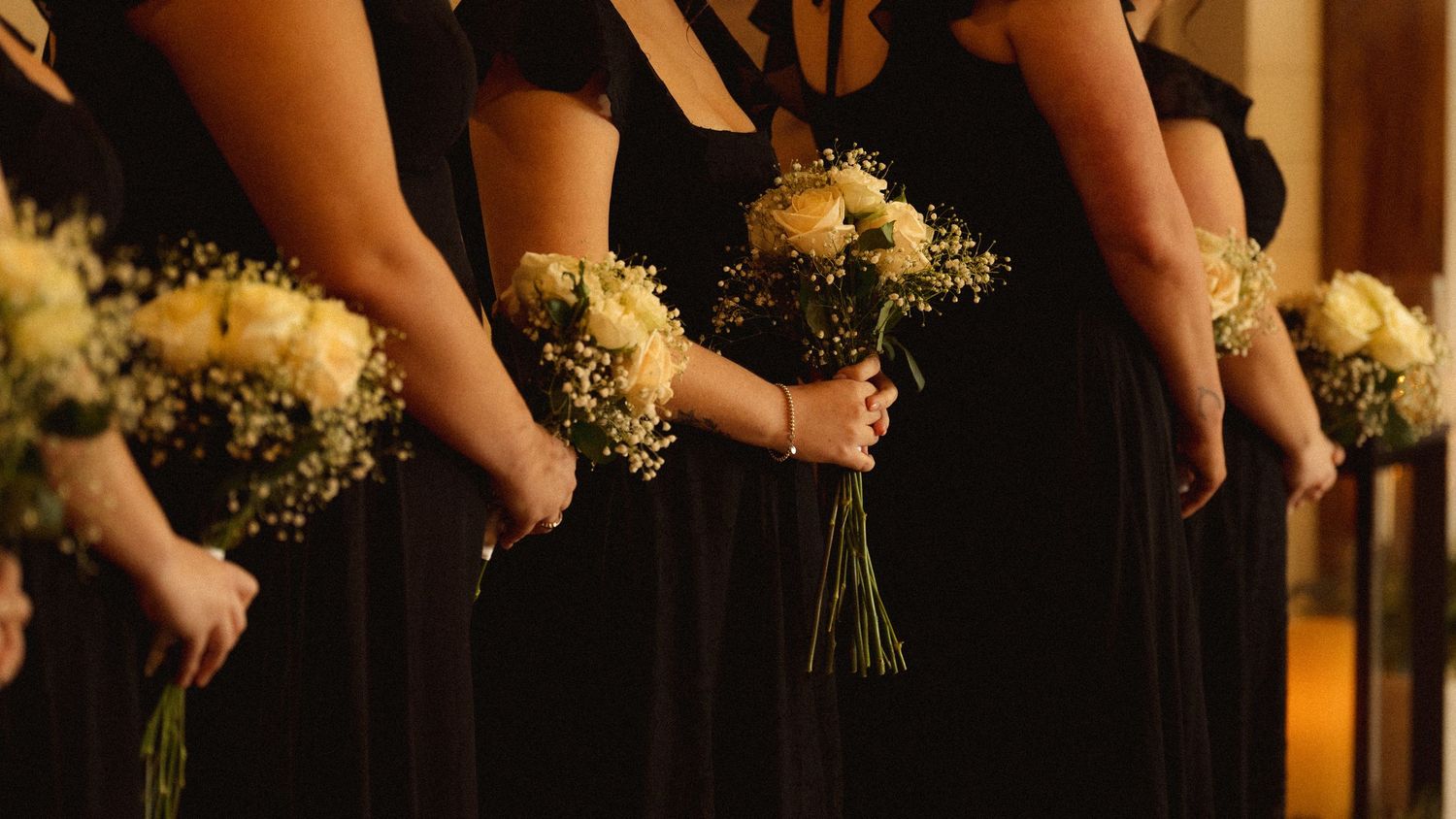 Wedding party holds bouquets of white and blush flowers in a row against a warm indoor backdrop.