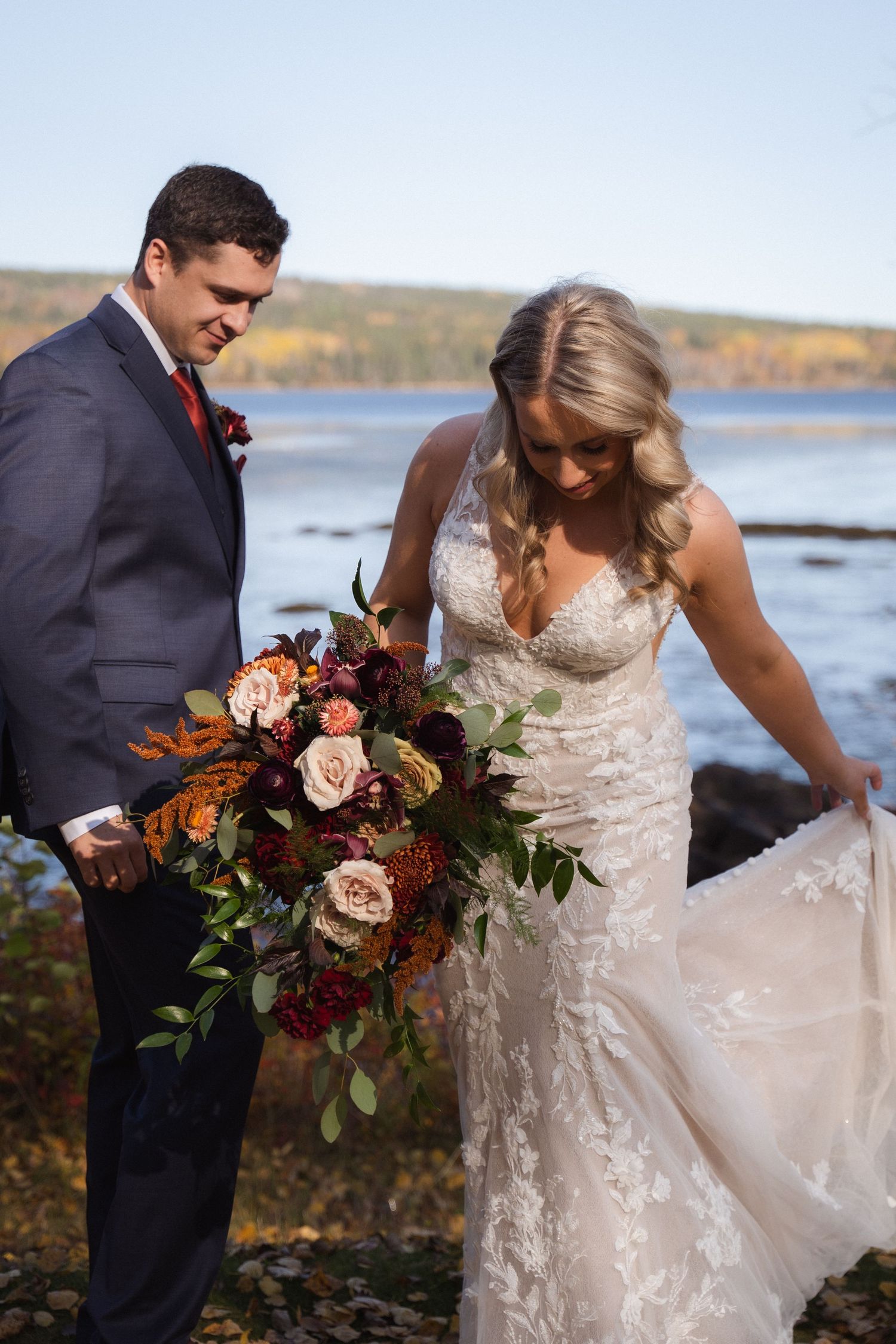 A bride in a lace wedding dress poses with her bouquet by a waterfront landscape.