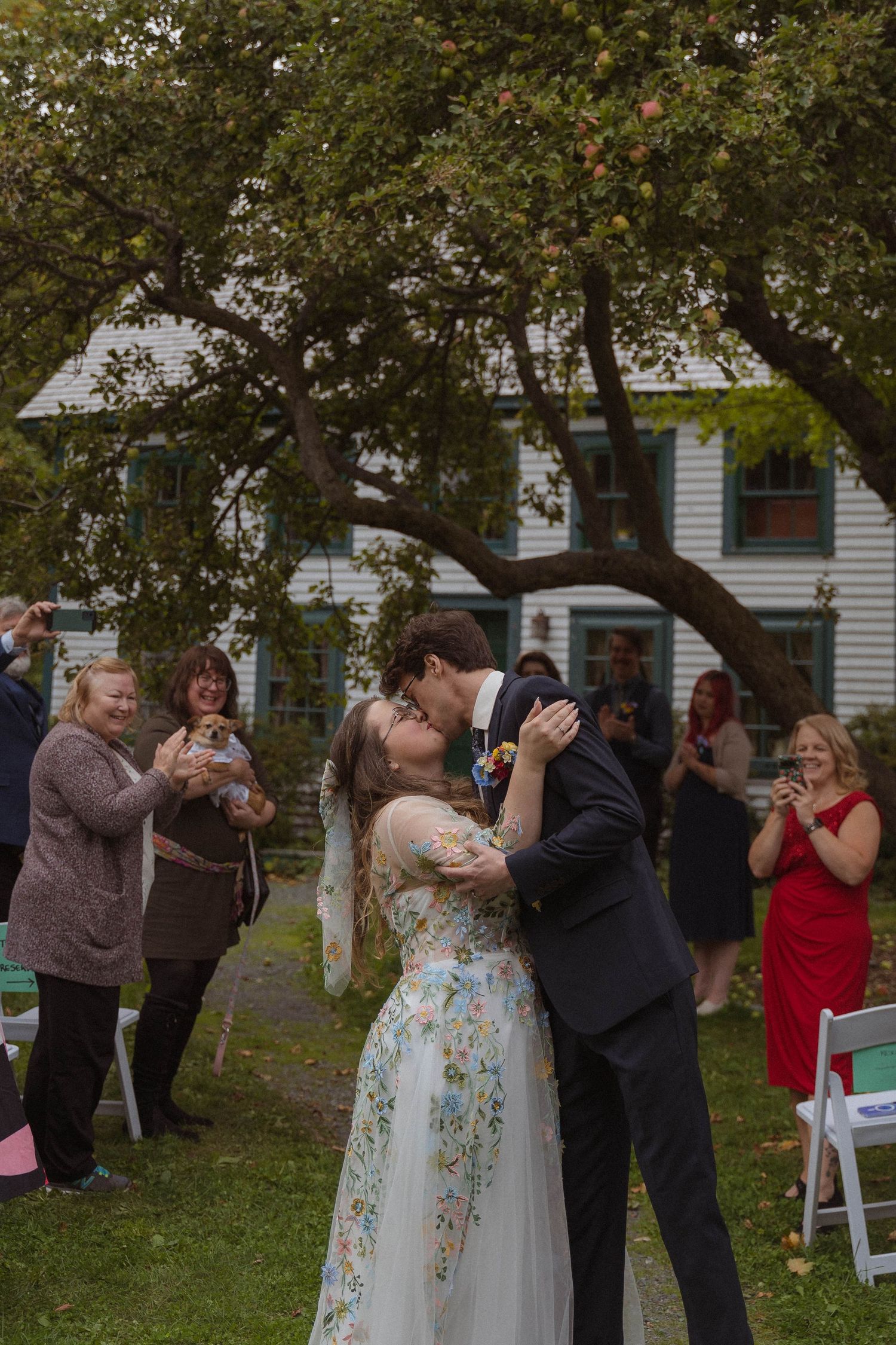 A couple shares their first kiss at an outdoor wedding ceremony while guests look on and take photos.