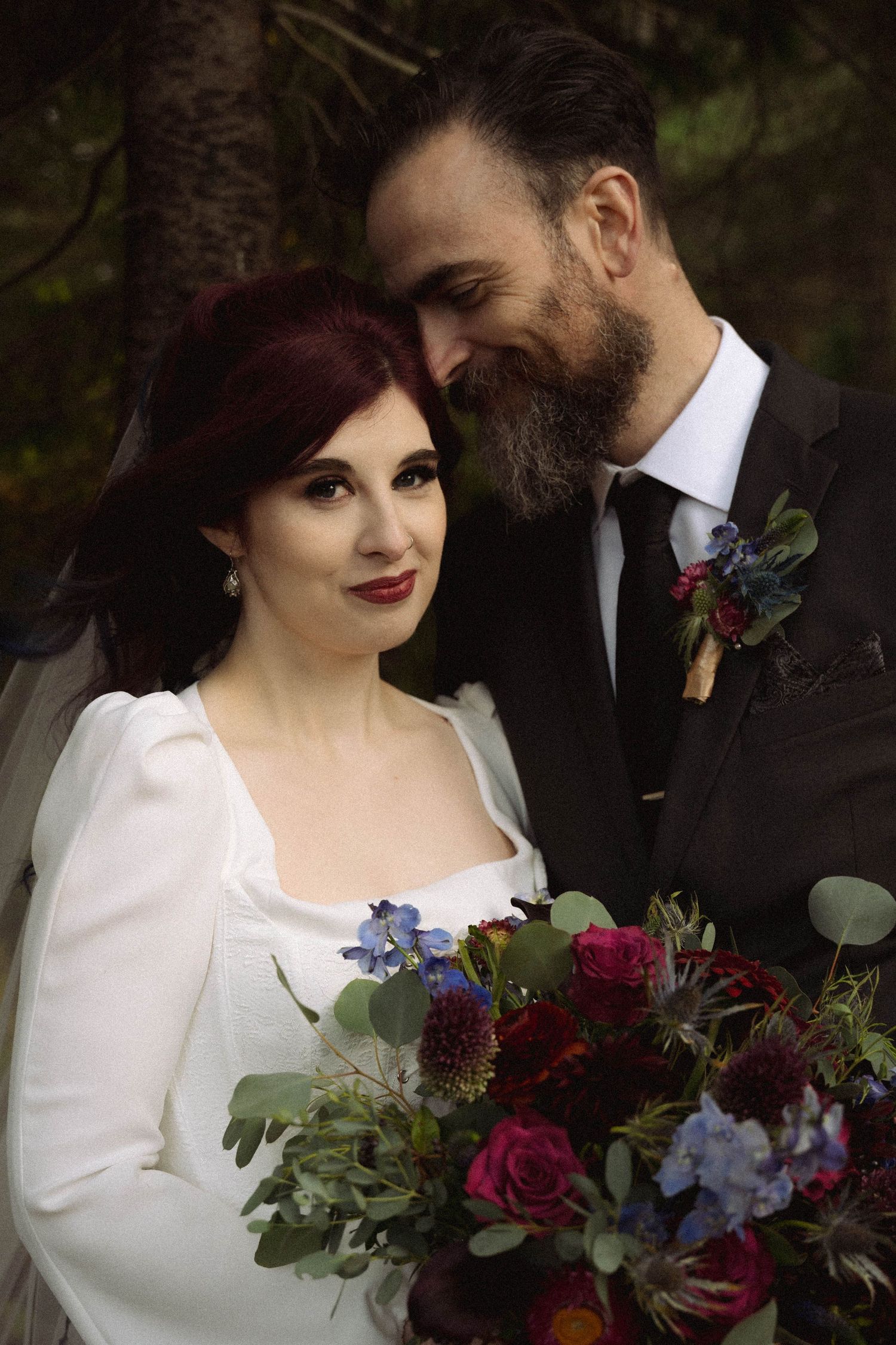 A romantic wedding couple poses in a garden setting with a stunning burgundy and purple floral bouquet.
