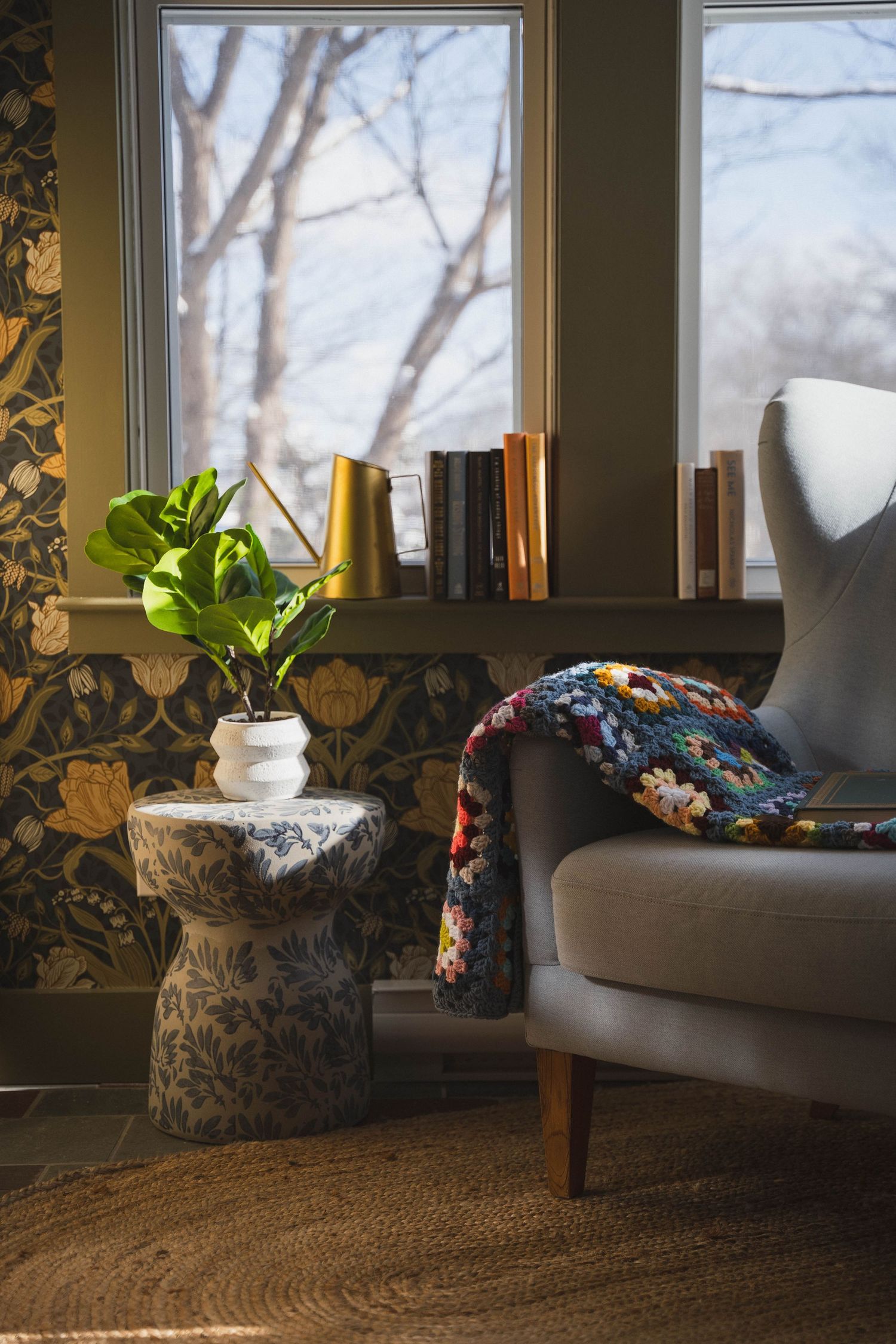 Cozy reading nook with gray wingback chair, colorful throw, botanical wallpaper, plant on side table, and books by sunny window.