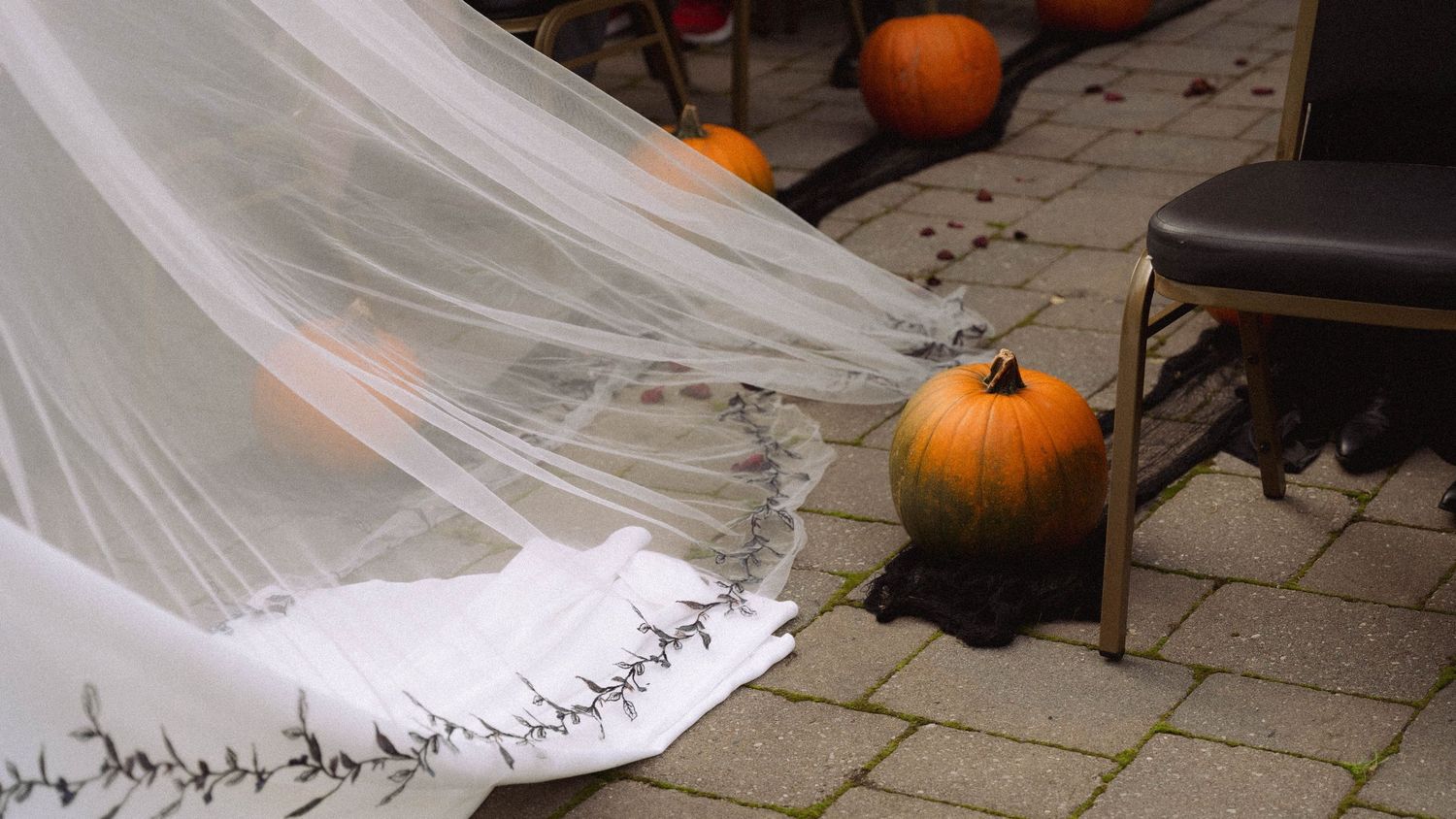 A flowing white wedding dress train and veil trail across stone pavement next to orange pumpkins.