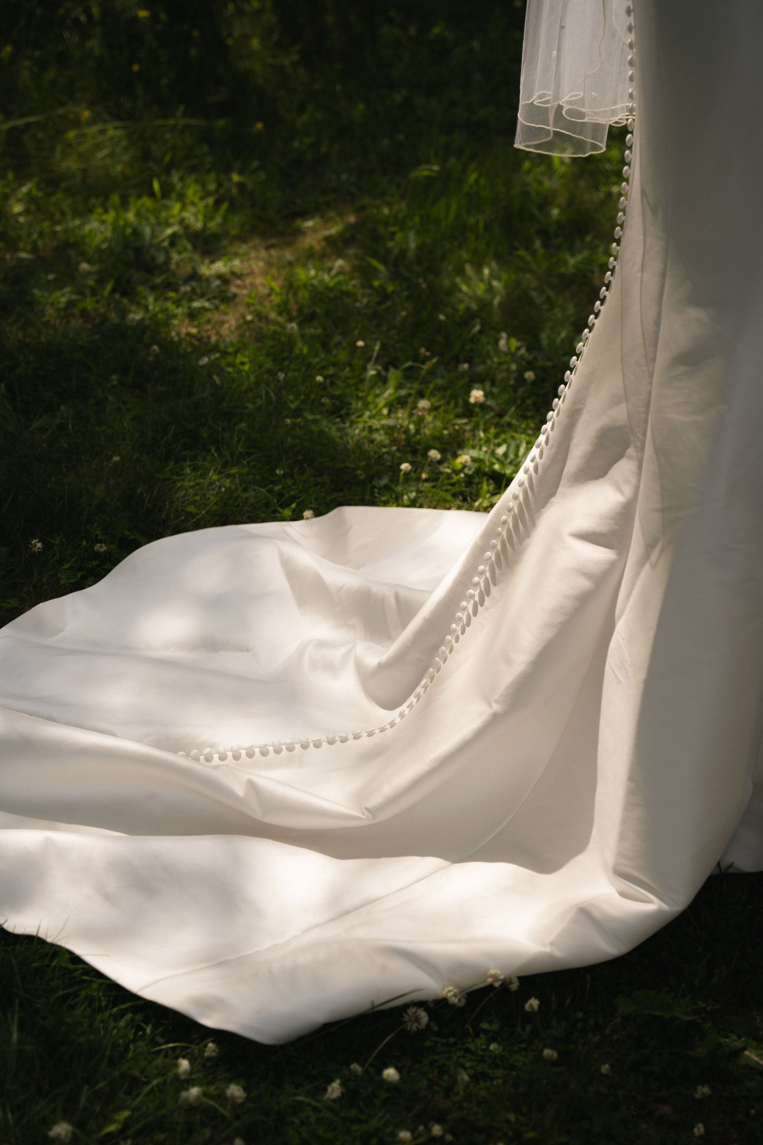 White wedding dress with delicate pom pom trim flowing in the breeze against green grass.